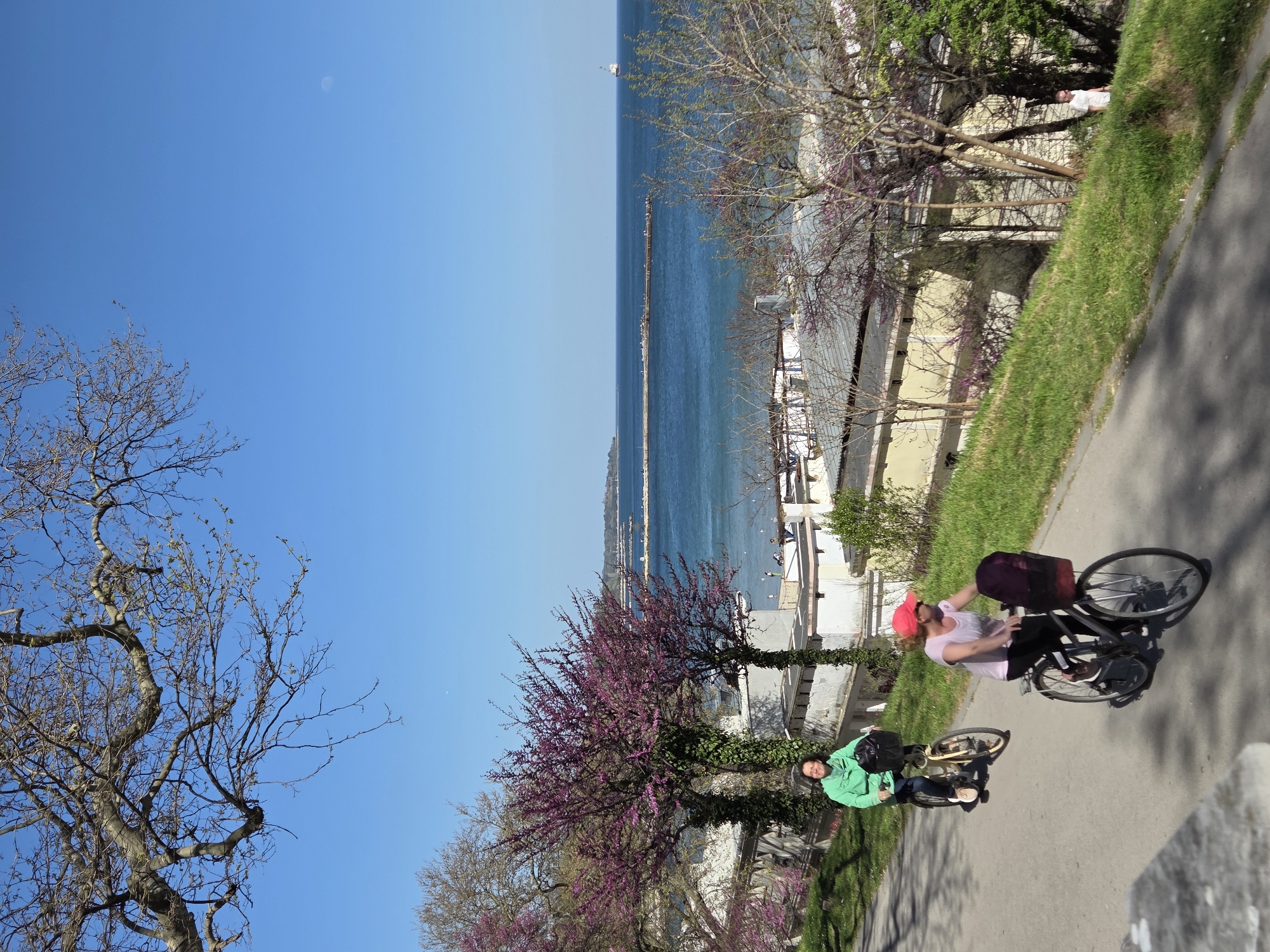 Two people are riding bicycles on a paved path along the Bulgarian Black Sea coast. The blue sea and sky are visible in the background. The location is in the city of Varna, Bulgaria.
The first person, on the right, is wearing a pink hat and a light-colored shirt. The second person, on the left, is wearing a green jacket and is on a smaller bicycle. Both people are on bikes with what looks like packages behind them.
There are trees and grass along the path. Buildings are also visible behind the trees.