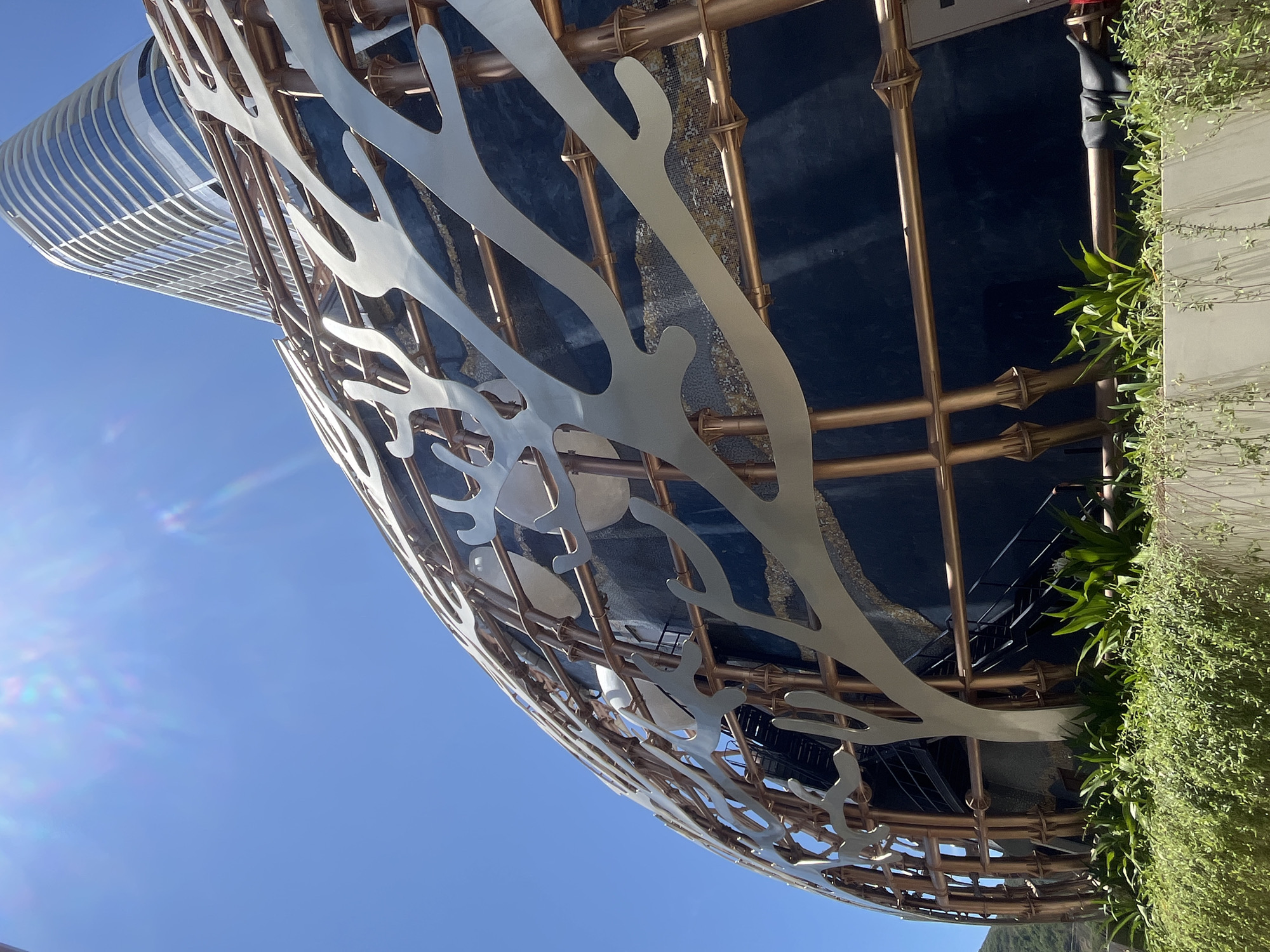 The image presents a low-angle view of an architectural structure against a clear blue sky, likely taken in Bac Nha Trang Ward, Vietnam, though specific location details within the ward are not discernible from the image alone. The structure features a complex, curvilinear design with a metal framework that is primarily gold in color. This framework is overlaid with flat, abstract, light-colored panels resembling organic shapes, possibly mimicking coral or branches.
In the background, another structure with a rounded, multi-story design and a facade of horizontal white lines is visible.
On the right side of the image, there's dense greenery with various plants and vines cascading down the side of the structure, suggesting the presence of a vertical garden or landscaped area.
The overall impression is of modern architecture intertwined with natural elements.