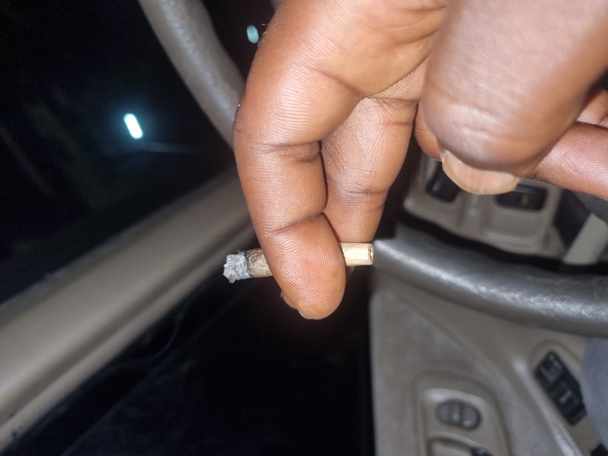A close-up shows a brown-skinned hand holding a lit cigarette or joint between the thumb and forefinger, inside a vehicle. The ashed end is visible. A steering wheel and dashboard elements are visible in the background. The location cannot be determined from the image, there is no evidence to suggest this is the city of Jos, Nigeria.