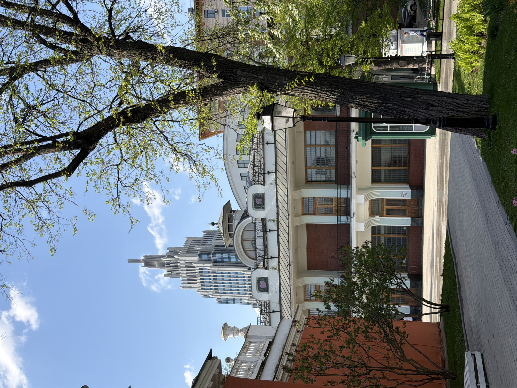 The image depicts an urban scene in Moscow, Russia, featuring a mix of historic and modern architecture. A large tree with light green foliage dominates the left side of the frame. In the background, a tall, modern skyscraper with a distinctive, stepped design rises against a blue sky with scattered clouds. A closer building, with a light-colored facade, appears to be a historic building with ornate detailing, arched windows, and decorative balconies. A street lamp stands near the tree. A paved walkway stretches along the right side of the image. A small strip of grass and some greenery are visible adjacent to the walkway.