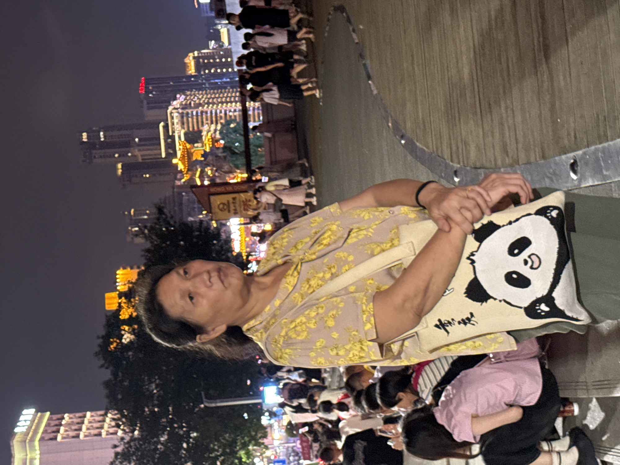 A woman with dark hair, wearing a yellow floral print shirt and green pants, stands in the foreground of a busy nighttime street scene in Yuzhong District, China. She is holding a beige tote bag with a panda design. In the background, numerous people are walking and standing around, with illuminated buildings and trees visible. The street appears to be a pedestrian walkway with a curved wooden boardwalk. The overall atmosphere suggests a lively urban environment at night.