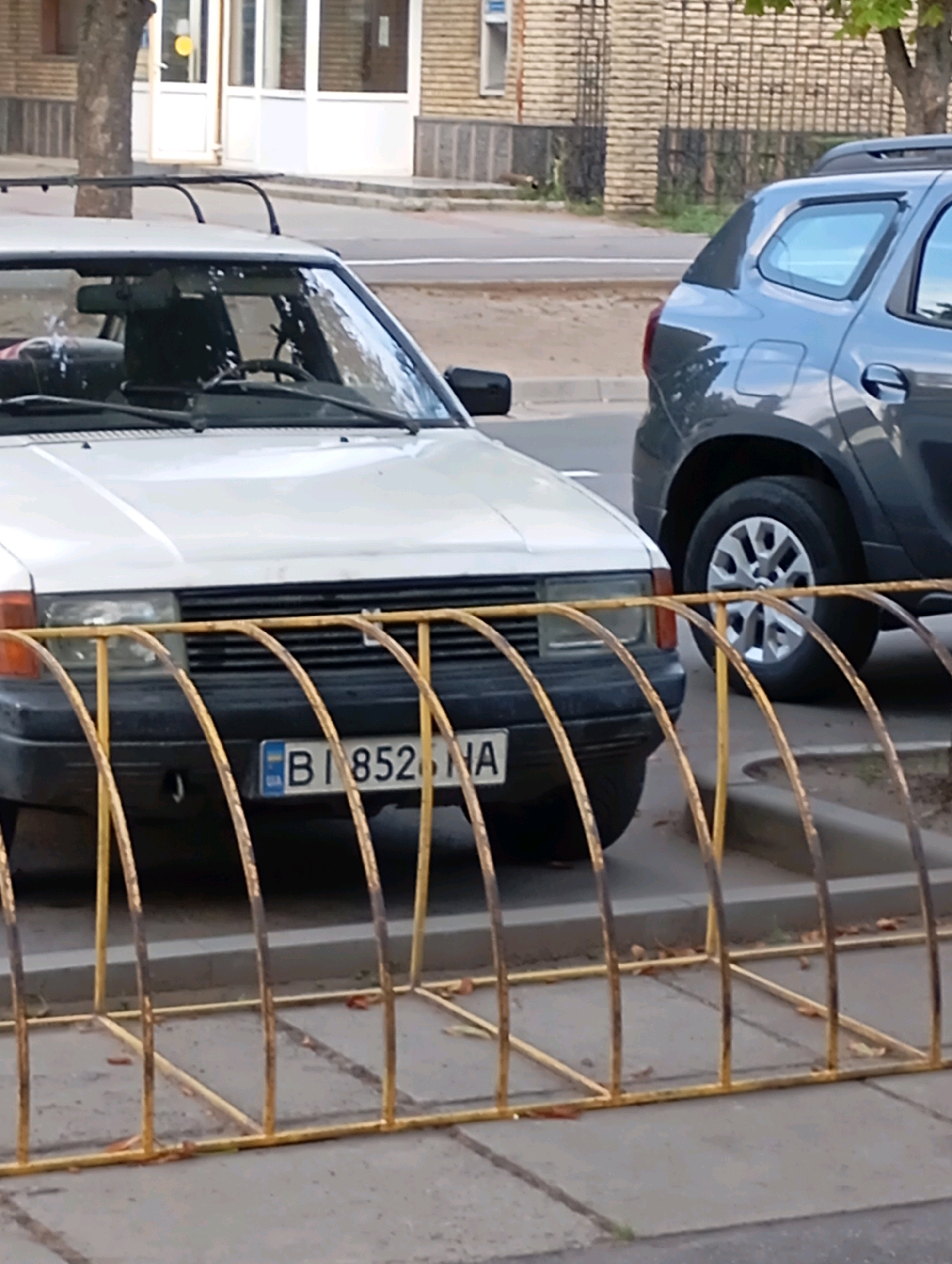 Two cars are parked on a street in Horishni Plavni, Ukraine. The car in the foreground is white, and its license plate is visible, reading "BI 852 NA". Behind it, a darker-colored SUV is parked. A yellow metal bike rack is situated in front of the white car. The street appears to be paved, with a sidewalk visible. Buildings and trees are present in the background.
