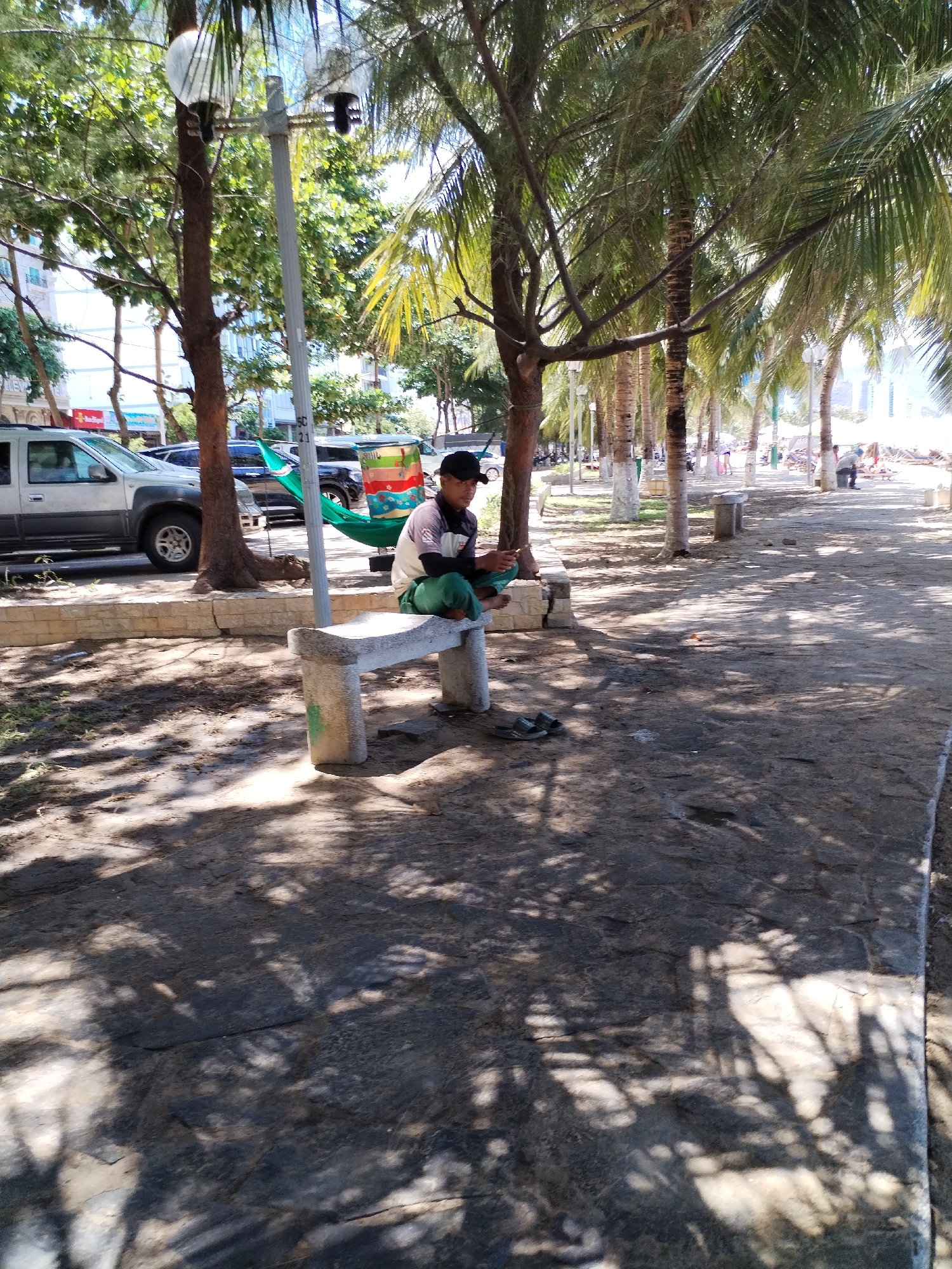 A man sits on a concrete bench in a park located in Bac Nha Trang Ward, Vietnam. He is wearing a black cap, a light-colored shirt, and green pants. The man appears to be looking at or using a handheld device. A pair of sandals sits on the ground near the bench.
Behind the bench, there's a parked silver SUV, a green hammock strung between two trees, and a refreshment cart. The park is lined with palm trees and other trees. In the background, people stroll along a path that leads to a beach. The ground is covered in a mix of stone and dirt, dappled with shadows from the trees.