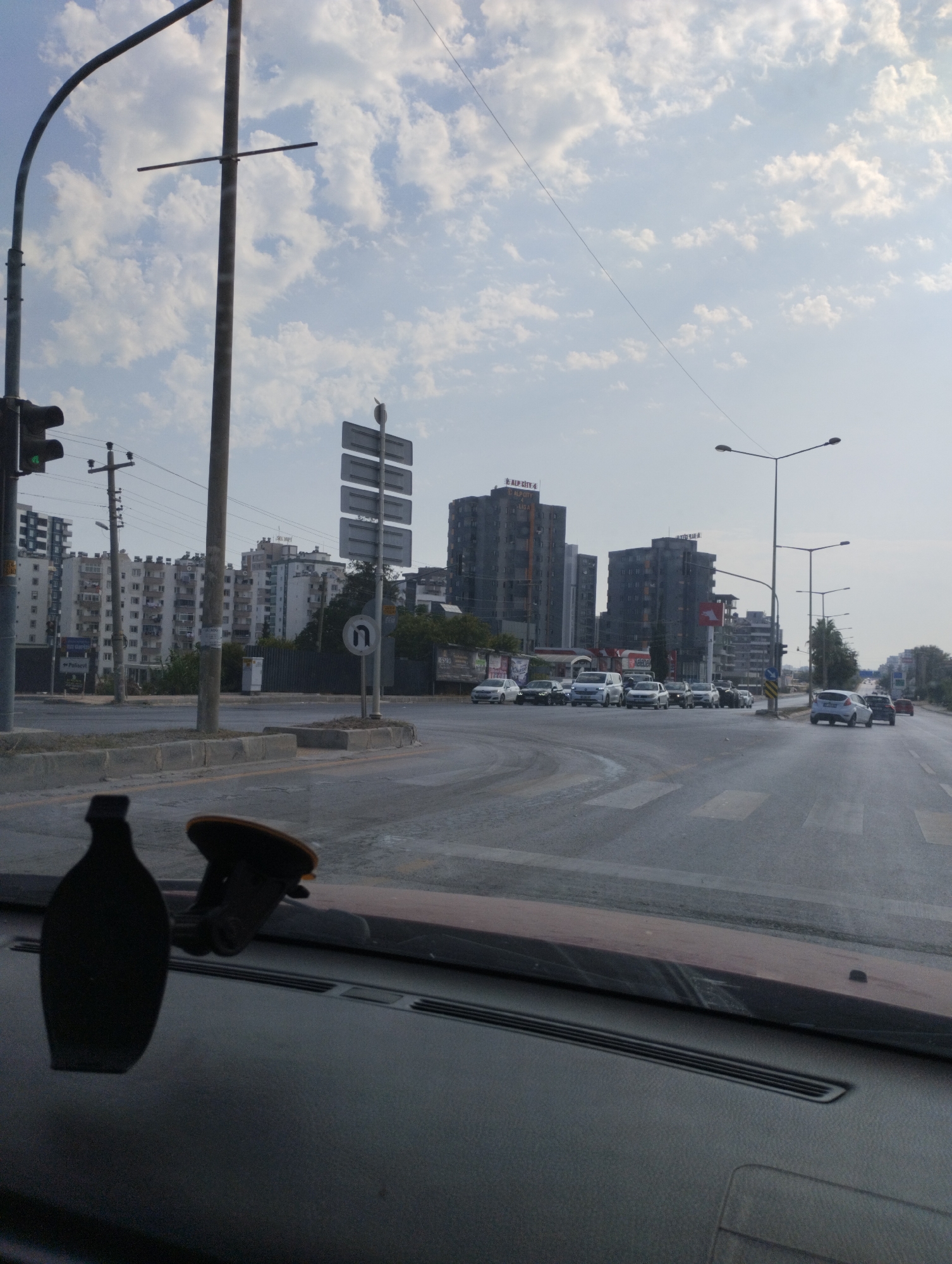 The image depicts a street scene in Mezitli, Turkey, under a partly cloudy sky. A car's dashboard with a black suction cup phone mount is visible in the foreground. The road ahead has crosswalk markings and several lanes of traffic. A variety of vehicles, including cars and vans, are present on the road. In the background, multiple apartment buildings and commercial structures stand. Traffic lights and street lamps are also visible. A sign indicating a U-turn is posted on the right side of the road. The overall environment suggests an urban setting with moderate traffic.