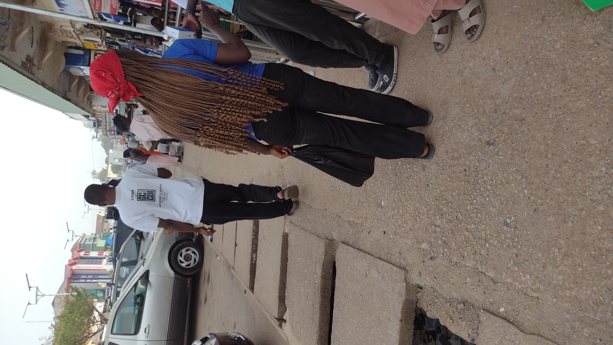 The image shows a street scene in Jos, Nigeria. A woman with long, light brown braids and a red headscarf is standing on the sidewalk. She is wearing a blue shirt and black pants and holding a black plastic bag. Next to her, another person is wearing a pink top and black pants. To the left, a man in a white t-shirt with a black design is walking near a parked silver van. The street is paved and the sidewalk is concrete with visible seams. Buildings and cars are visible in the background. The sky is overcast.