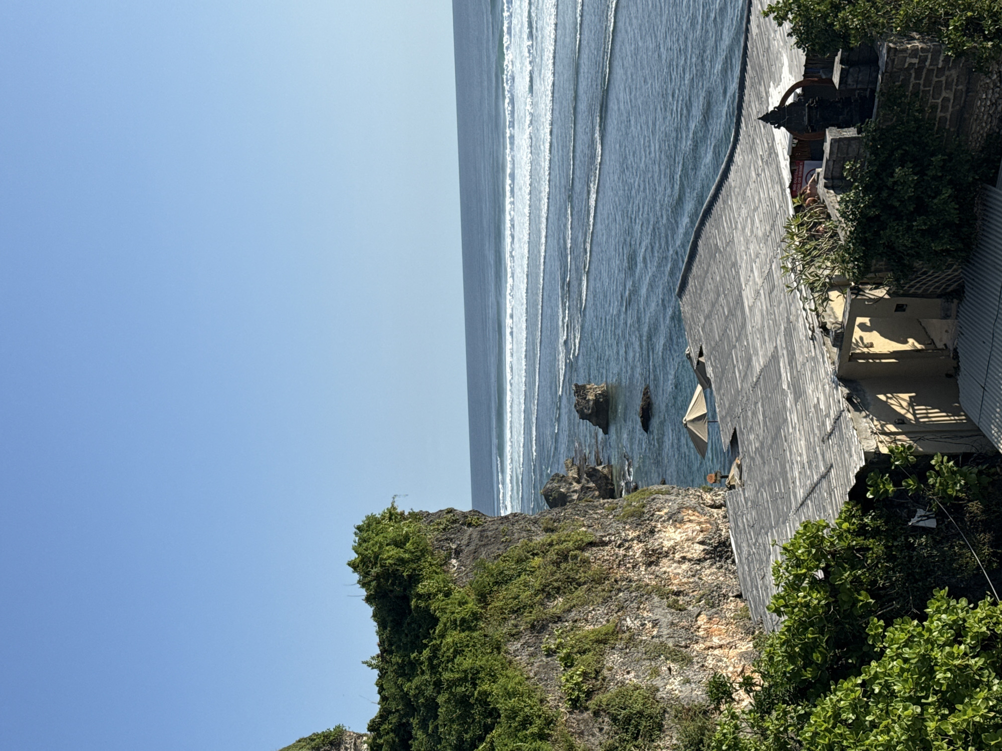 The image shows a coastal scene in Kuta Selatan, Indonesia. A rocky cliff covered in vegetation dominates the foreground. The ocean stretches out with visible wave patterns and some rock formations near the shore. To the right of the cliff, a building with a wooden exterior is visible. The sky is clear and blue.