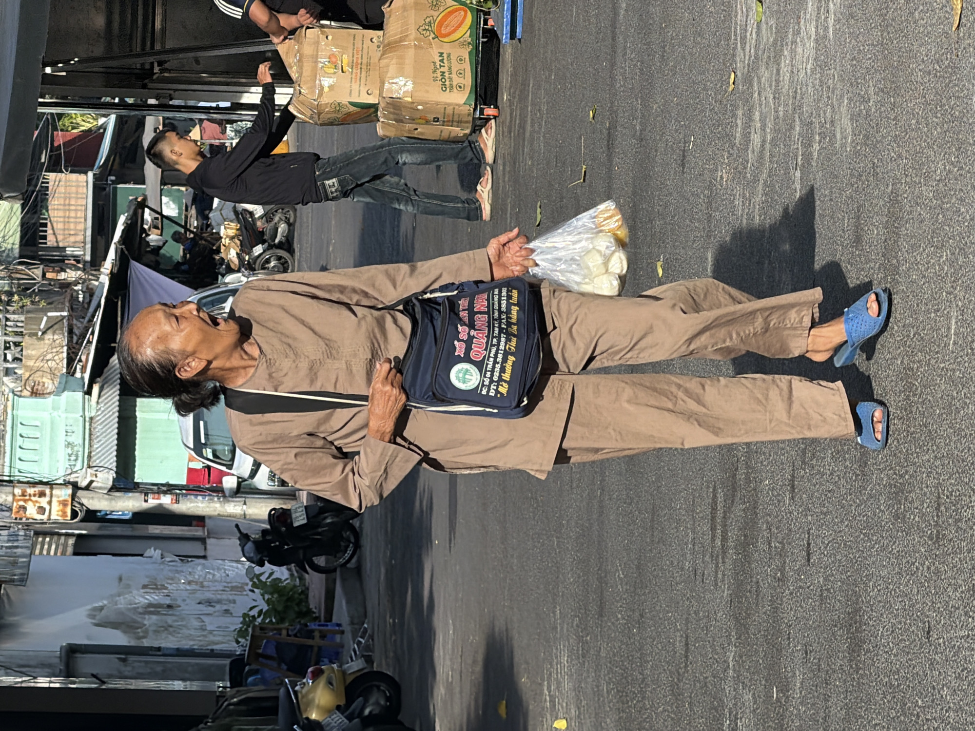 The image shows a street scene in Nha Trang, Vietnam. An elderly person in loose-fitting brown clothing, with their mouth open as if speaking or yawning, walks towards the right. They are carrying a blue shoulder bag with text and graphics, and holding a clear plastic bag containing what appear to be food items. The person is wearing blue slip-on sandals. Behind them, a younger person in a black long-sleeved shirt and jeans stands near stacked cardboard boxes. The environment appears to be a marketplace or street with buildings, a parked motorcycle, and sunlight casting shadows.