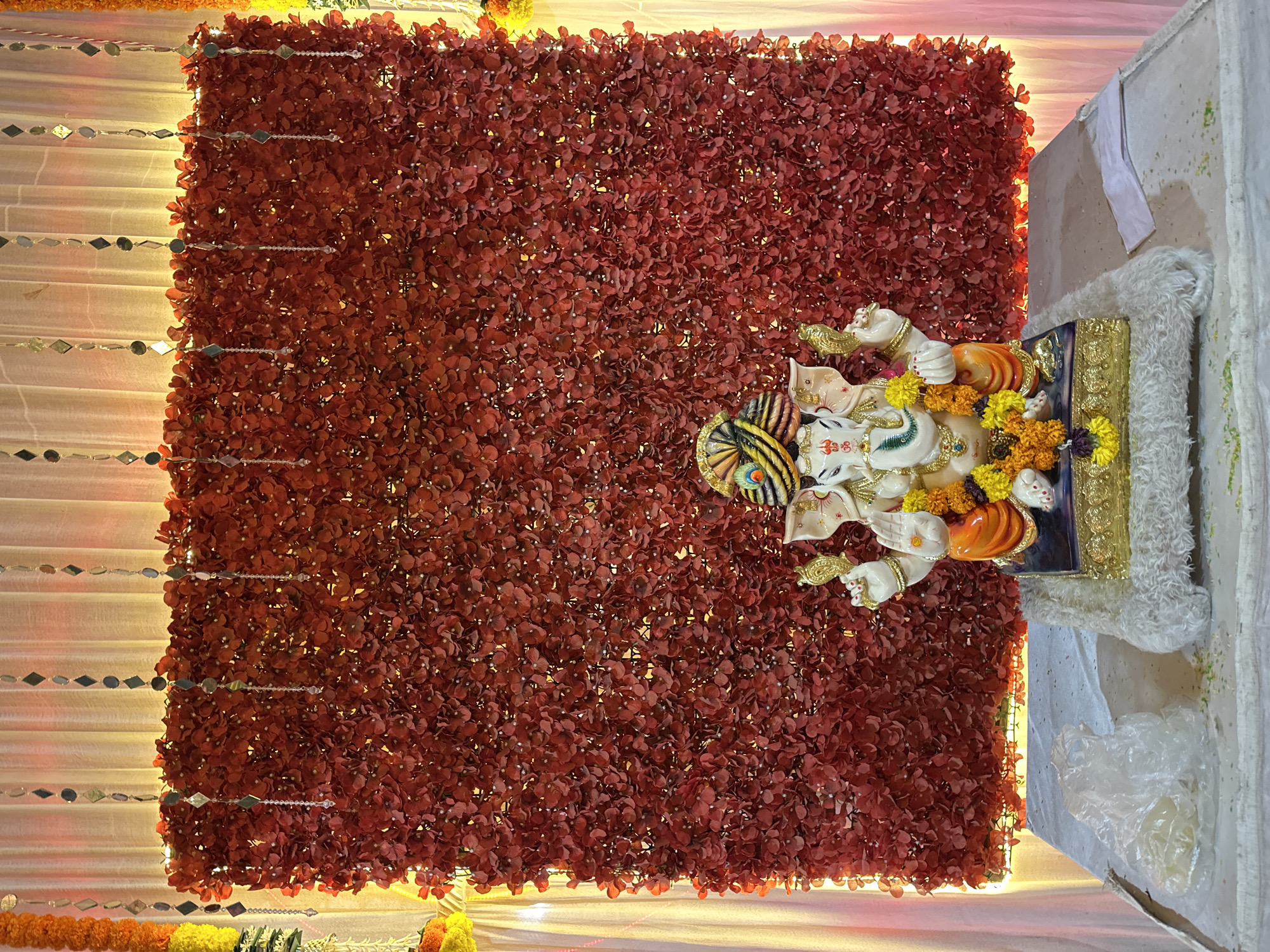 The image shows a statue of Ganesha placed in front of a backdrop of red flowers. The statue is adorned with yellow and orange decorations. To the left of the flower backdrop, there is a draped white fabric decorated with strings of small mirrors. The scene appears to be an indoor setting, possibly for a religious event or celebration. While the location is Shahapur, India, specific details about the precise venue or event are not provided.
