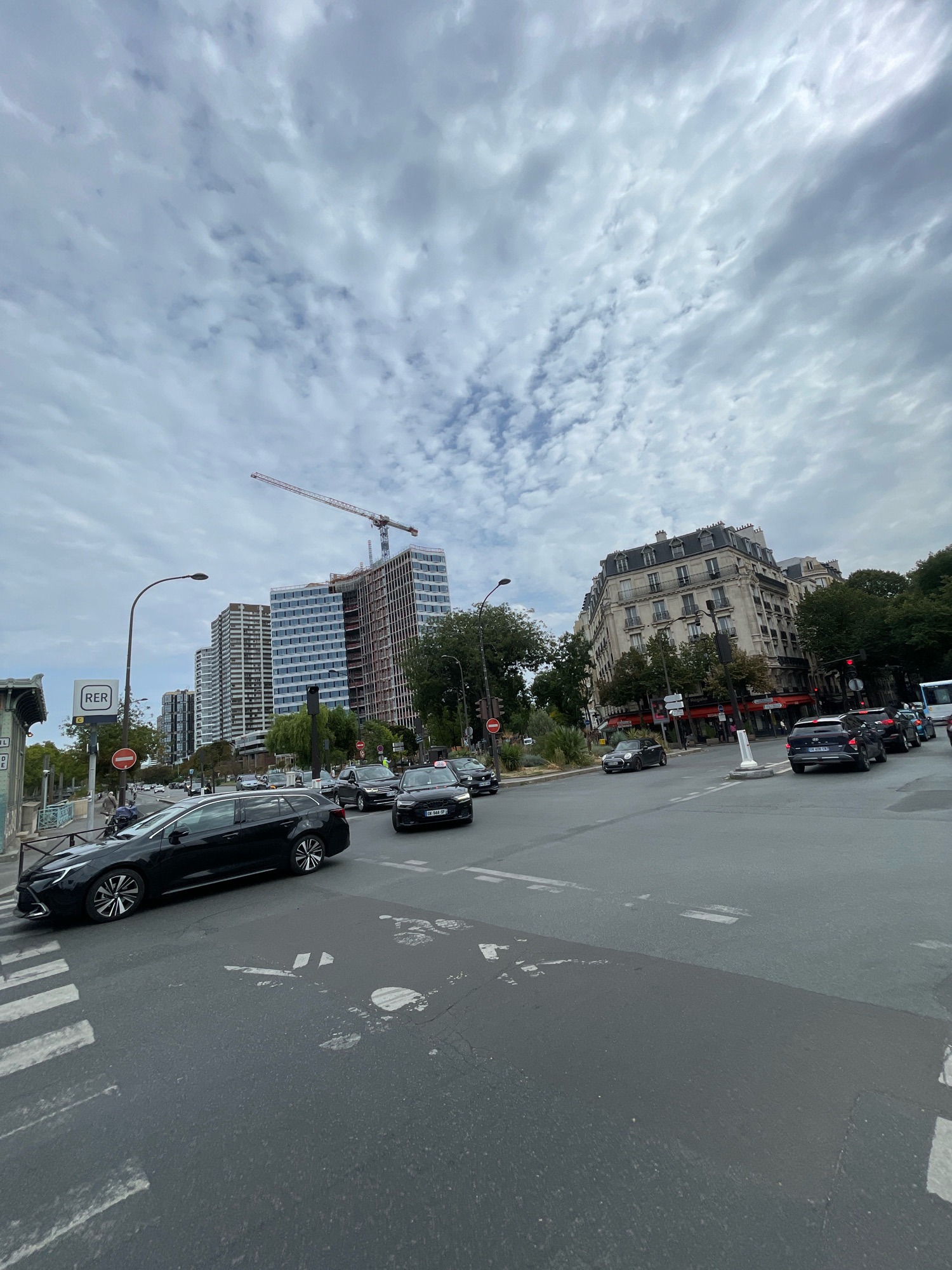 The image captures a street scene in Paris, France, on a cloudy day. In the foreground, a black station wagon is parked on the left side of a pedestrian crossing, with its front wheel slightly over the white lines. The road has various lane markings, including white dashed lines and a painted bicycle symbol.
Further down the street, traffic is flowing. Several cars, including black sedans and SUVs, are visible on the road. One car has a "Taxi" sign on its roof. To the left of the road, a lamppost stands next to a sign indicating "RER," a regional train network in Paris. Further left, there's a glimpse of a green building with arched entrances.
In the background, a modern, tall building under construction is a prominent feature, with a red construction crane extending from its top. Adjacent to this construction site are several other tall, contemporary buildings with glass facades. To the right, an older, ornate Parisian building with multiple stories, balconies, and a mansard roof stands. Trees with green foliage line the street, and traffic lights are visible. The sky is filled with a mix of white and gray clouds.