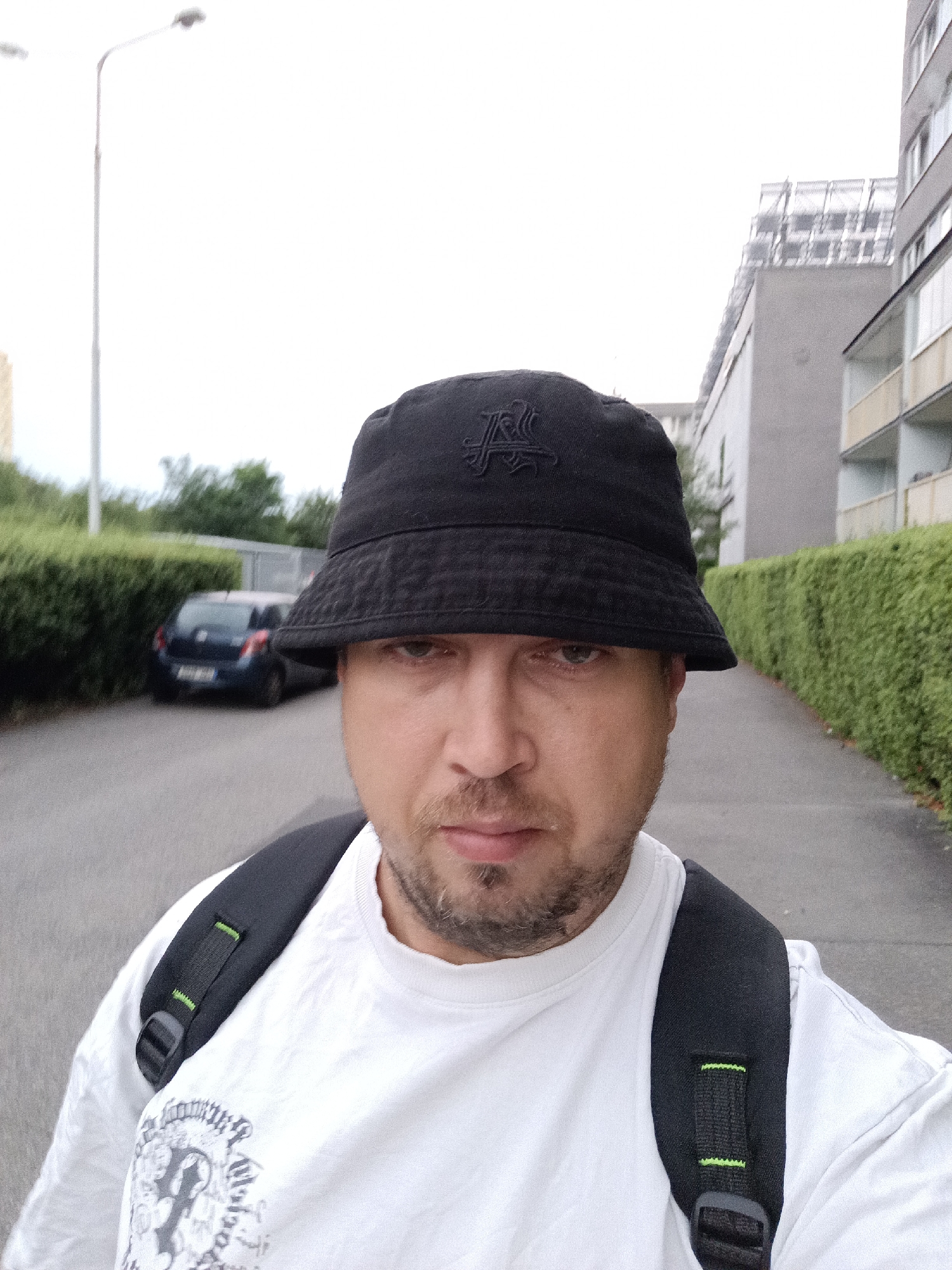 A man with a beard and a black bucket hat is captured in a close-up, low-angle selfie. He is wearing a white t-shirt with a graphic design and a black backpack. The man is outdoors, standing on an asphalt path. In the background, a dark blue car is parked on the left side of the path, with a manicured green hedge running alongside it. Further in the background, apartment buildings and a street light are visible under a cloudy sky. The scene is likely a residential area in Prague, Czechia, given the architectural style of the buildings and the presence of street lighting typical of European cities.