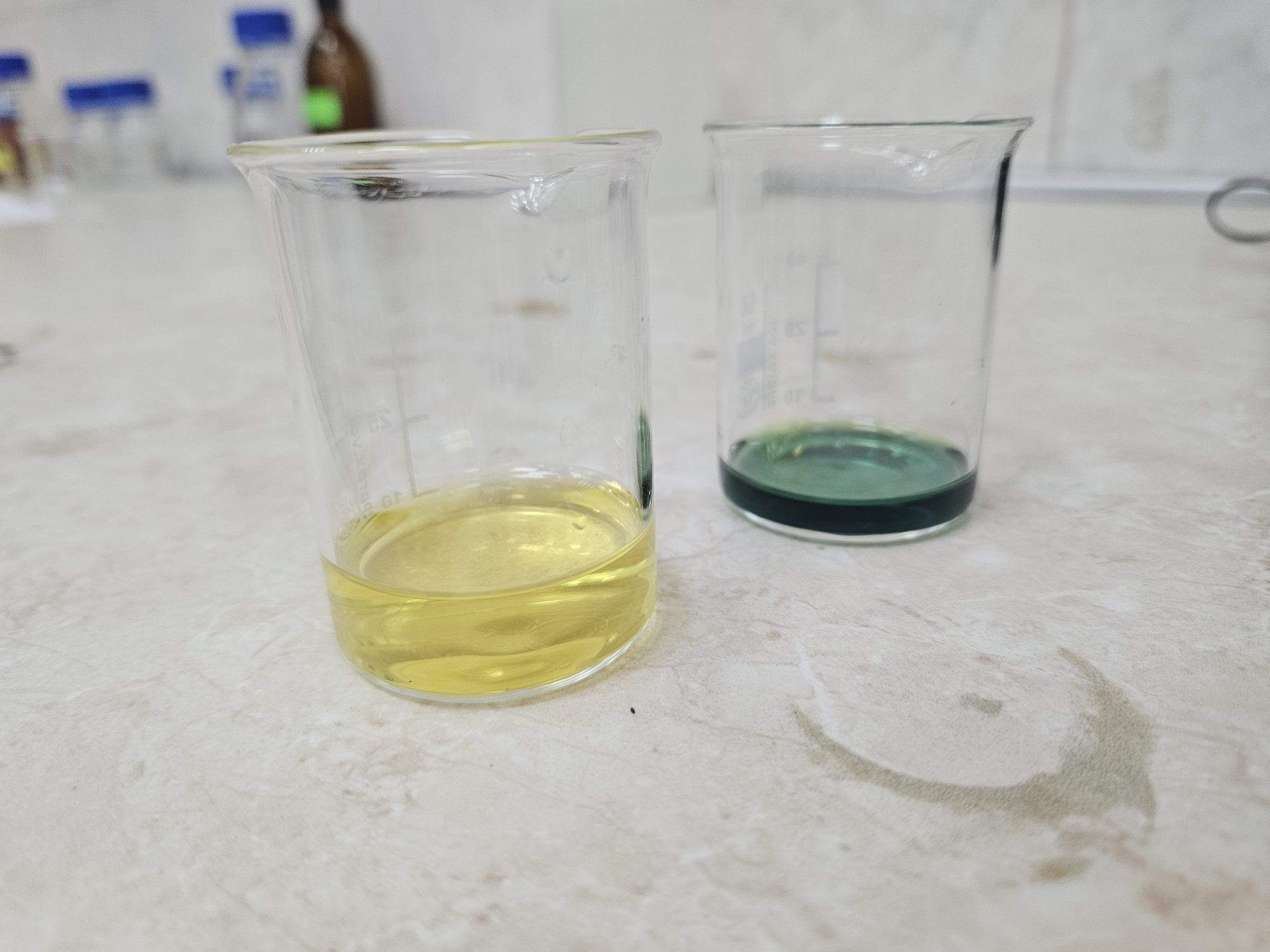 Two beakers are on a counter. One beaker contains a yellow liquid. The other contains a green liquid. The background shows lab equipment. The location is unknown. No people are visible. No actions are visible.