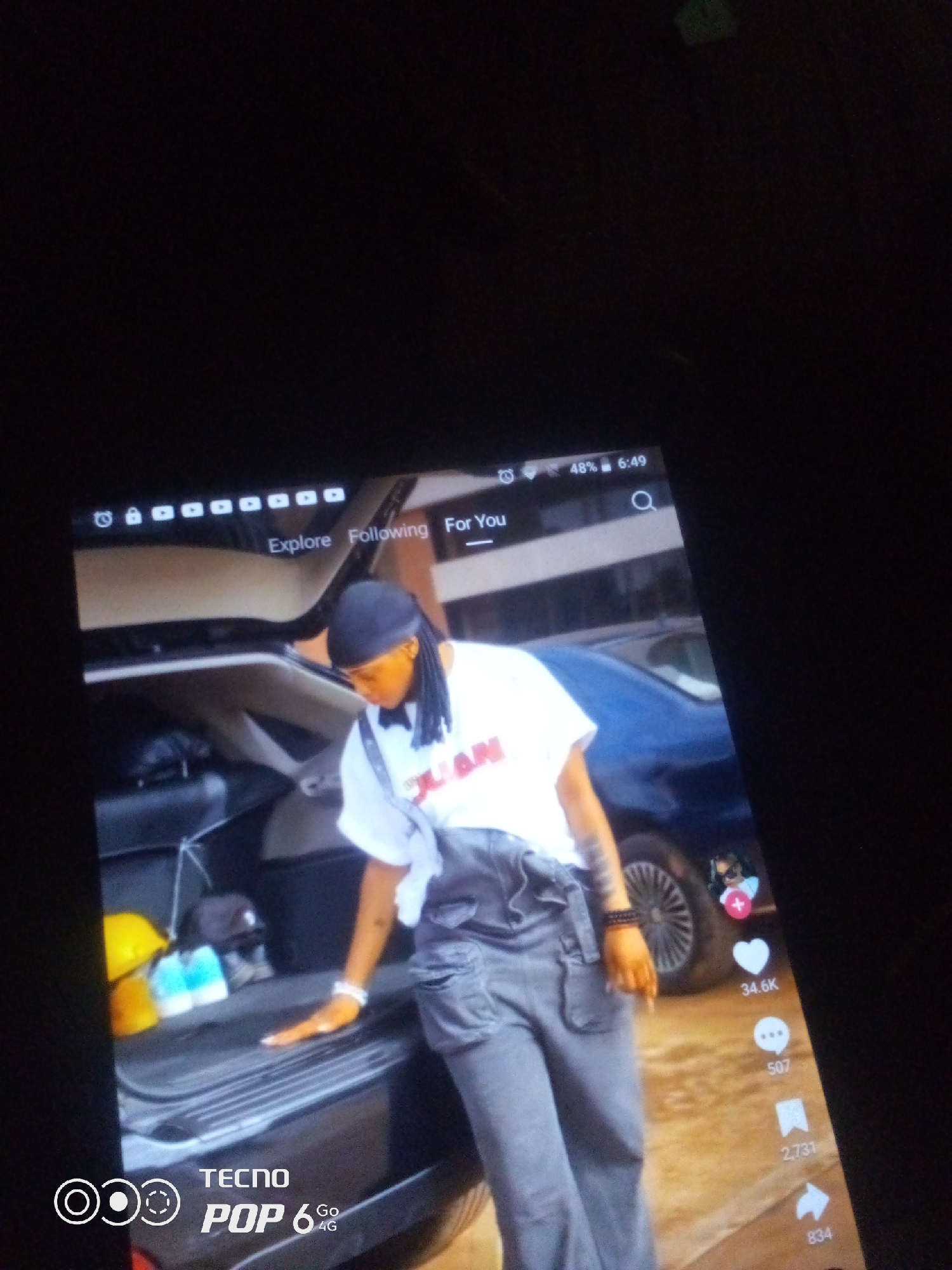 A person is partially visible in the image, wearing a white t-shirt with red lettering and gray overalls. They have on a black durag and bracelets on their wrist. The person is standing next to the open trunk of a car, which contains two yellow hard hats and two blue and white bottles. The background shows a blue car and a dirt or gravel surface. A tablet displaying a social media feed is also visible, with icons for likes, comments, shares, and other engagement metrics. The top of the tablet shows the time as 6:49 AM and the battery level at 48%. The image is taken from an angle, with the tablet screen dominating the frame. The location of Howayi, Nigeria is not depicted in this image.