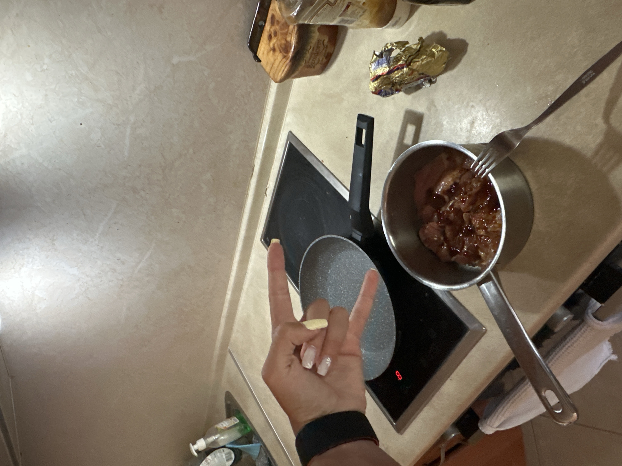 A close-up, overhead shot captures a kitchen counter in Botevgrad, Bulgaria, featuring elements of food preparation. A person's hand, adorned with white and yellow-tipped fingernails and a black fitness band on the wrist, is positioned above a stovetop, forming a "rock on" gesture. In the foreground, a silver pot contains food coated in a dark sauce, and a fork rests beside it. To the right, a piece of crinkled gold foil and a glass jar are visible. A small, light-colored ceramic object with a wooden lid sits on the counter. The background shows a beige tiled wall and a stovetop with a red indicator light.