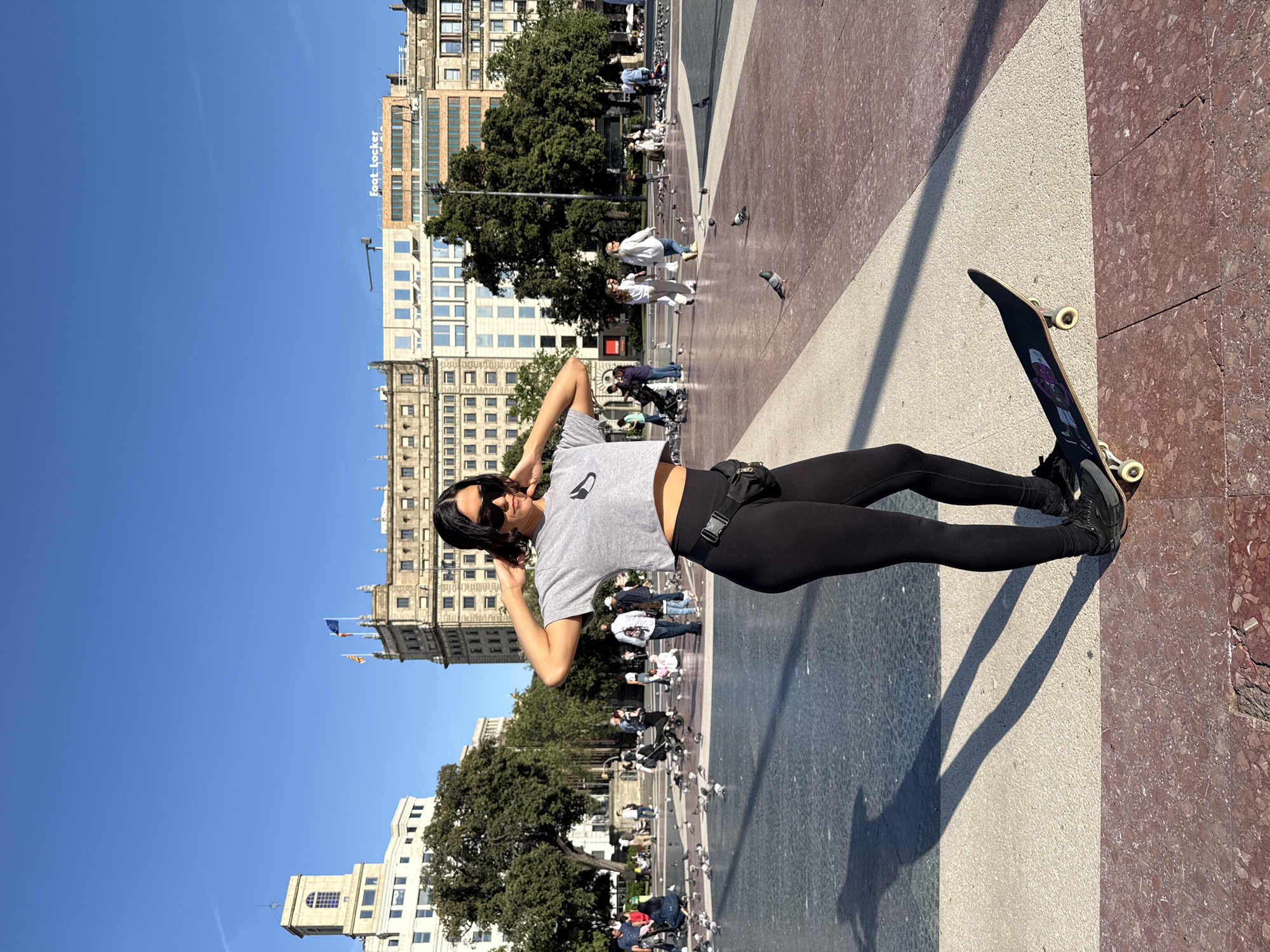 The image depicts a woman in black leggings and a gray t-shirt standing in Plaça de Catalunya, a large square in central Barcelona, Spain. She has dark hair and sunglasses and is posing with her hands behind her head. Beside her is a skateboard. The square is paved with a mosaic of colors and patterns. In the background are buildings, trees, and a crowd of people with pigeons. A building with "foot-locker" is visible. The sky is clear and blue.