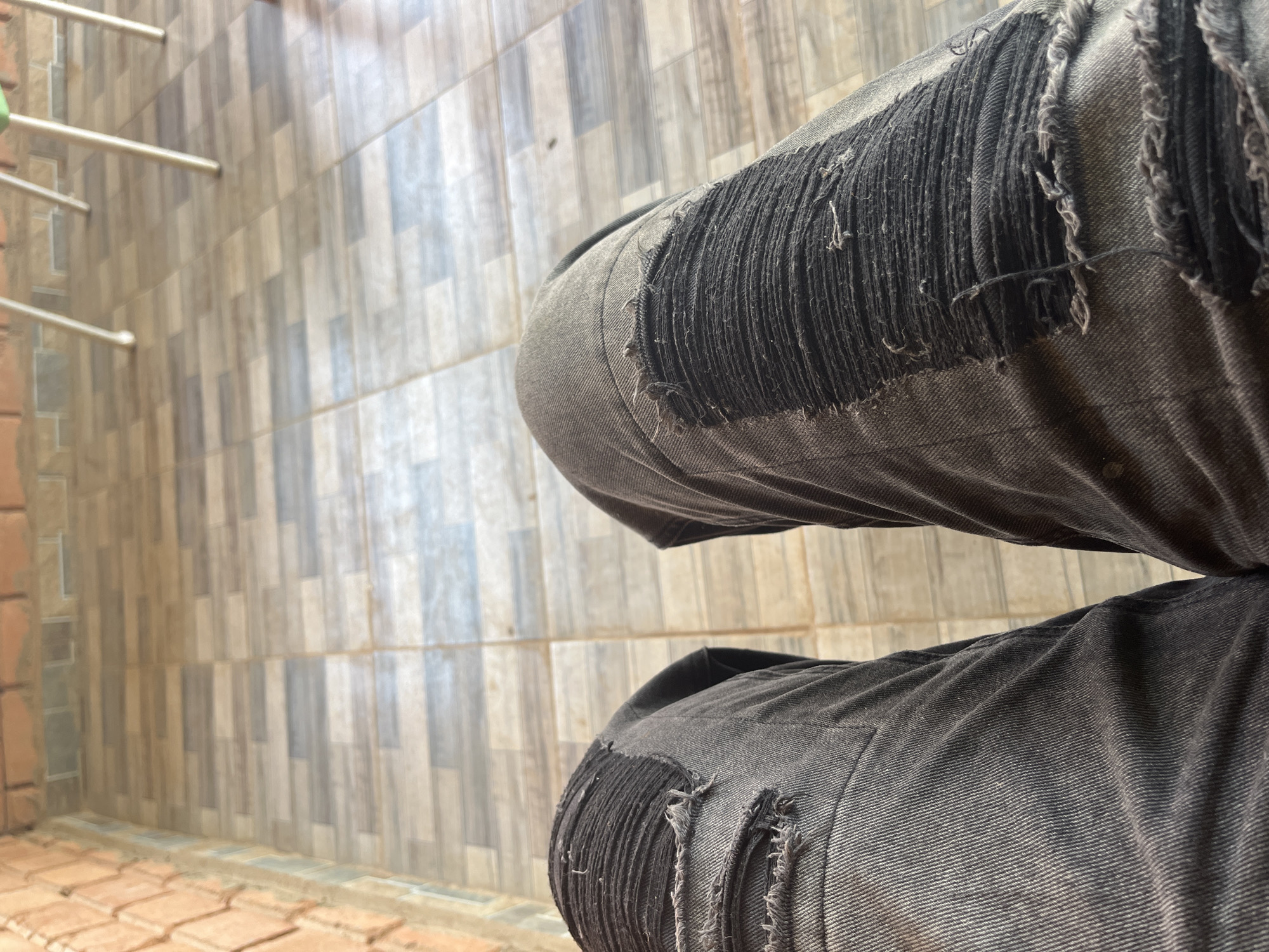 The image shows a close-up view of a person's legs wearing ripped black jeans. The background is a tiled floor with a brick wall on the left. The scene is indoors or on a covered patio, possibly in Jos, Nigeria, given the commonality of tiled floors and brick construction in the region. There are no people visible other than the legs, and the focus is on the texture and detail of the jeans and the surrounding floor. The lighting appears natural, suggesting it's daytime.