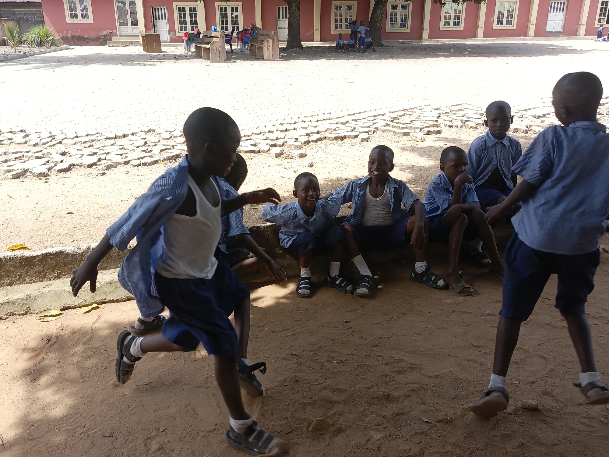 This image depicts a vibrant outdoor scene featuring a group of approximately ten young boys, likely students, in what appears to be a schoolyard or courtyard in Akwanga, Nigeria. The setting is bright and sunny, indicated by strong shadows, suggesting midday or early afternoon.
The boys, who are Black, are dressed in similar blue striped shirts, dark shorts, white socks, and sandals, characteristic of a school uniform. In the foreground, two boys are in motion, suggesting active play; one is running towards the left, while another stands with his back to the viewer, appearing engaged in the activity. The other boys are gathered under the partial shade of trees in the midground, some sitting on the sandy ground and others on low wooden benches. Several of these seated boys are smiling broadly, conveying a sense of happiness and camaraderie.
The background features a two-story building with reddish-pink walls, multiple white-framed windows, and white doors. In front of the building, the ground consists of loose dirt and an area with broken, uneven paving stones. Stacked plastic chairs in red, blue, and green, along with more wooden benches, are visible near the building, suggesting an outdoor gathering space. The overall impression is one of relaxed playtime and community in a warm, informal school environment. No visible text is present in the image.