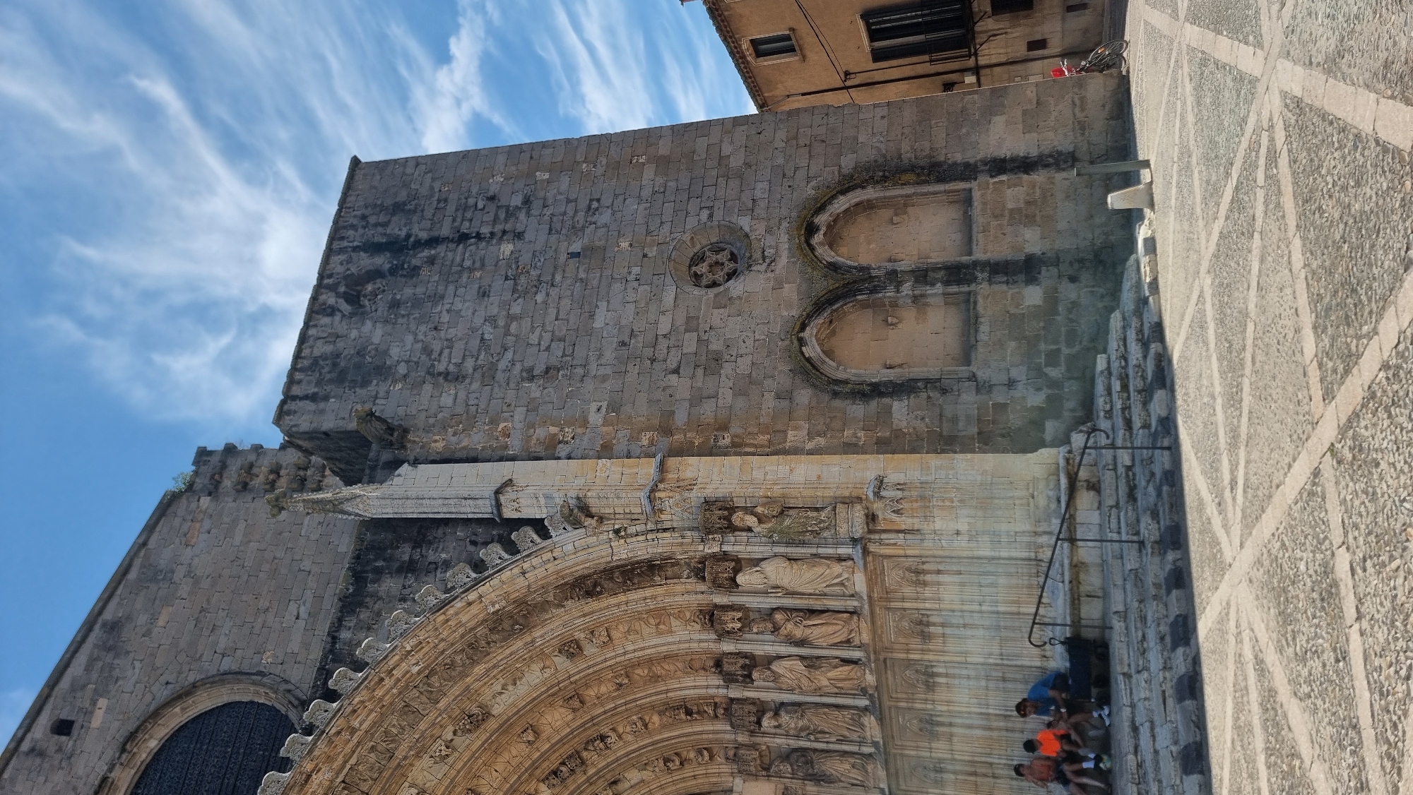 The image shows the exterior of the Basilica of Santa Maria de Castelló d'Empúries, a historic church located in the town of Castelló d'Empúries, Spain. The facade features intricate gothic architecture, including a large arched doorway with statues and decorative carvings. To the right of the entrance, a large stone tower rises, showing weathered stone blocks and architectural details like arched recesses and a circular window. In the foreground, a paved plaza with a cobblestone pattern and light-colored stone inlays is visible. Several people are seated on the steps leading up to the basilica entrance. One person is visible on a bicycle to the far right of the image. The sky above is blue with scattered wispy white clouds.