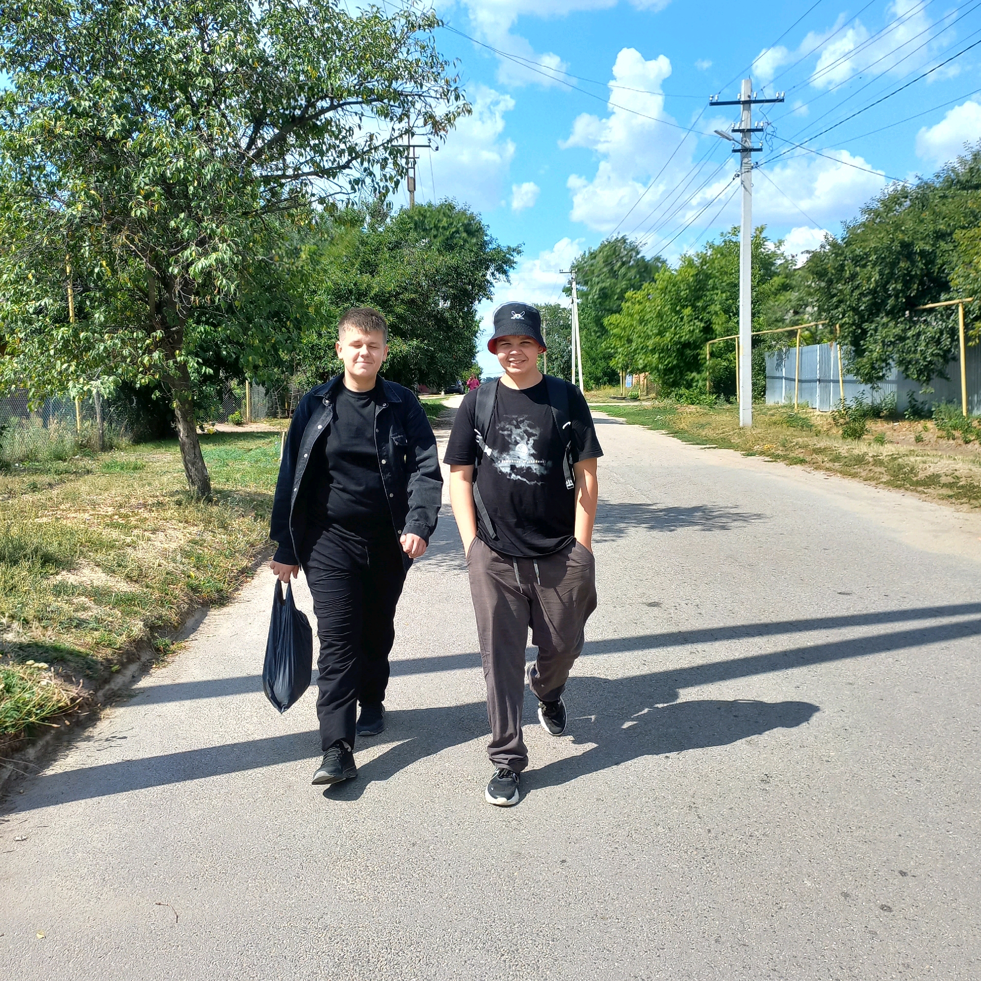 Two young males are walking down a paved road. The male on the left is wearing a black jacket, black t-shirt, and black pants. He is carrying a black bag in his left hand. The male on the right is wearing a black bucket hat, a black t-shirt with a white graphic, and gray pants. He has his hands in his pockets and a backpack on. They appear to be walking away from the camera, down a road lined with trees and a fence on the right side. The sky is blue with scattered white clouds. There are power lines visible overhead and a utility pole on the right. The lighting suggests it is daytime, with shadows cast on the road. The location is identified as Klyntsi, Ukraine.