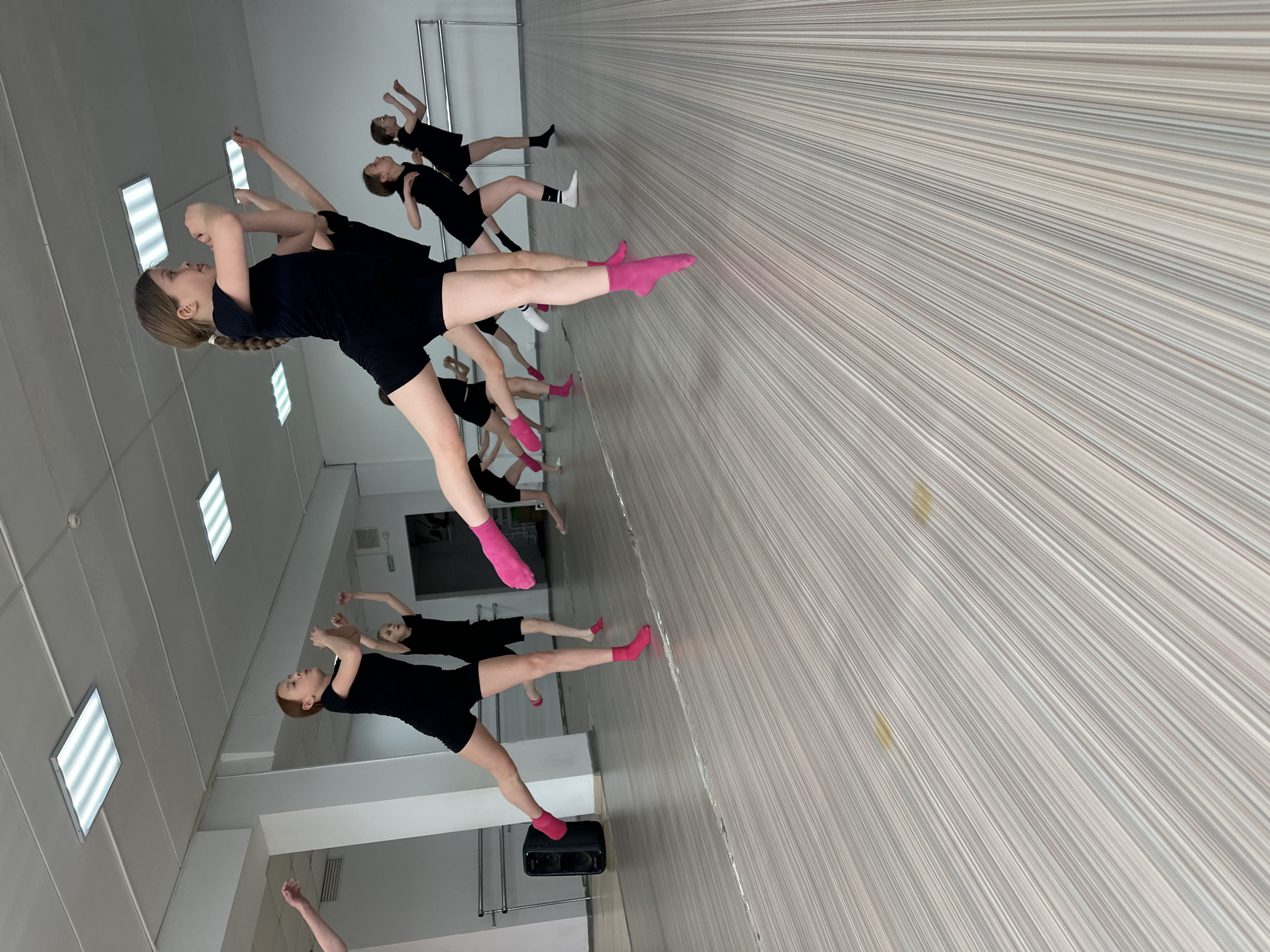 In an indoor dance studio, a group of young girls are engaged in a dance class. They are dressed in black leotards and pink or white socks. The studio is equipped with mirrors along one wall and a ballet barre on another. The walls are decorated with a beige and white striped wallpaper, and the floor is a light-colored wood. The lighting is provided by fluorescent ceiling lights. A black speaker is visible in the lower left corner. The girls are practicing a dance routine, with their arms raised and legs extended in various poses. The location is implied to be in Votkinsk, Russia.