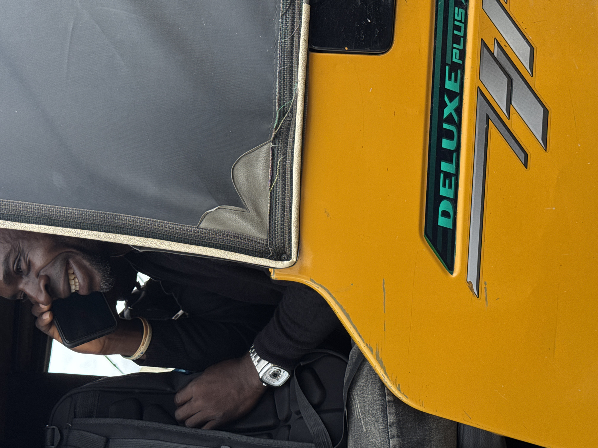A medium shot captures a man inside a "Deluxe Plus" auto rickshaw, likely in Ado, Nigeria. The man, dressed in black, is holding a smartphone to his ear, a wide smile revealing his teeth. A silver watch adorns his wrist, resting on a black backpack on his lap. The rickshaw's bright yellow exterior is marked with scratches, and the "Deluxe Plus" logo is prominently displayed. The interior features a dark grey fabric. The orientation of the photo is slightly rotated.