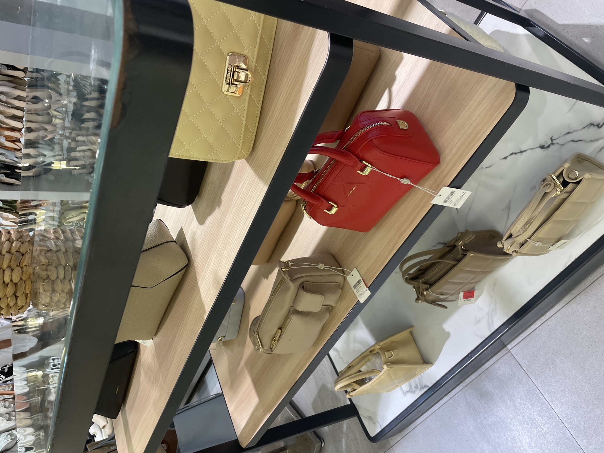 The image shows a display of purses inside of a retail store. The display is comprised of shelves with a black metal frame. The shelves on the left side are made of wood, and the shelves on the right side are made of marble. On the shelves are purses of different colors, shapes, and sizes. The purses are beige, red, and brown. The store is located in an unknown city.