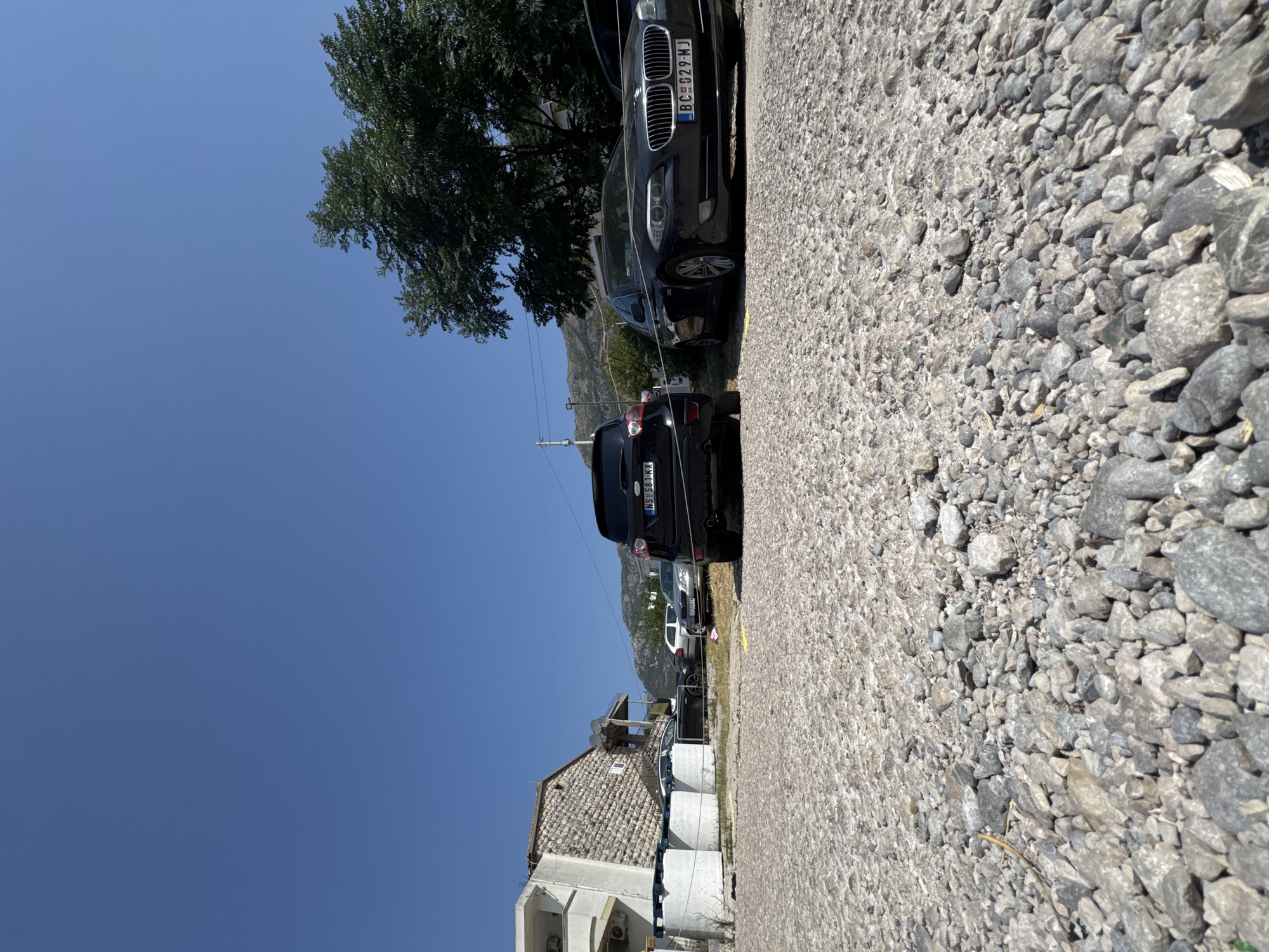 In the city of Sutomore, Montenegro, a low-angle shot captures a parking lot filled with gravel under a clear blue sky. The foreground is dominated by the textured gravel, with a few larger stones scattered amongst smaller ones. In the mid-ground, several parked cars are visible. To the left, a white building with stone accents and several large cylindrical containers sits near the parking area. Further back, a line of trees and rolling hills form the backdrop. A dark-colored SUV is parked on the right, its rear facing the camera. Adjacent to it, a dark sedan is also parked. The scene depicts an outdoor parking area on a sunny day.