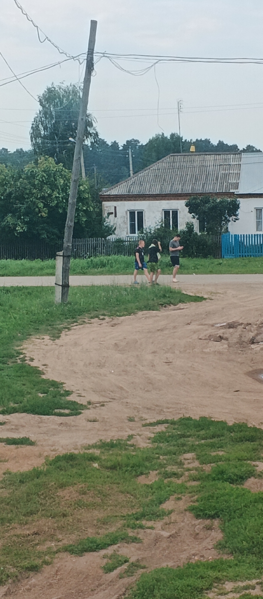 Three individuals, two young males and one young adult male, are walking on a dirt path in a rural area. The two younger males are walking close together, one appearing to be carrying something green attached to his back. The adult male is walking slightly ahead and to the right, looking down at his phone. They are walking away from the viewer, towards a white building with a corrugated roof in the background. The building has two windows visible and is surrounded by greenery. A wooden fence and a blue fence are also visible. A utility pole with wires is present to the left of the frame. The ground is a mix of dirt and grass. The sky is overcast. The scene is set in the village of Podgornoye, Russia.