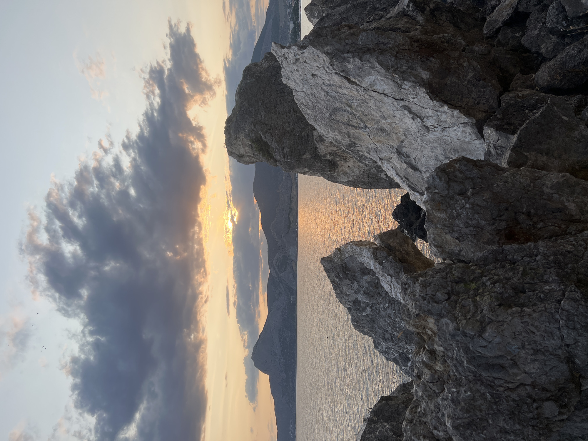 This image captures a dramatic sunset over the Black Sea near Sudak, Ukraine. In the foreground, large, jagged grey and white rocks dominate the scene, creating a rugged, natural barrier. The sea stretches out beyond the rocks, its surface shimmering with the golden hues of the setting sun. In the distance, a range of imposing mountains forms a silhouette against the sky. The sky itself is a canvas of soft blues and greys, with streaks of orange and yellow where the sun is setting, partially obscured by dark, heavy clouds. A few small birds are visible in the upper left portion of the sky. The overall impression is one of serene, natural beauty and a sense of remote wilderness.