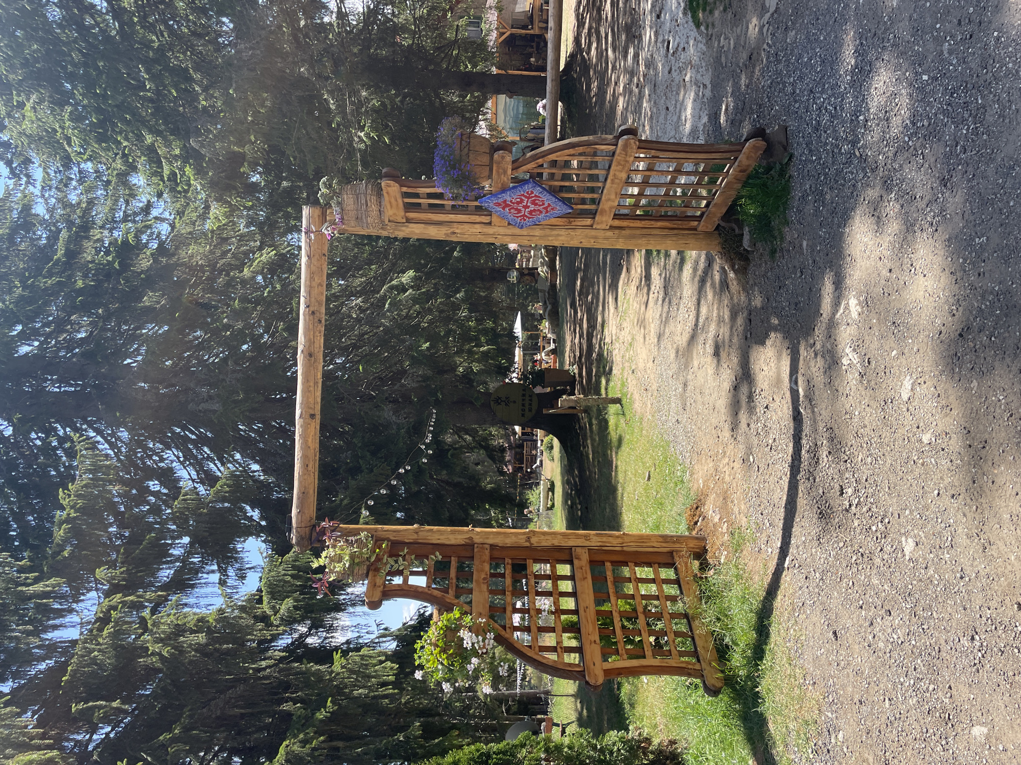 This is a vertical, eye-level, medium shot of a rustic wooden archway entrance to an outdoor area. The archway is constructed from unvarnished logs and features ornate, lattice-like wooden gates on both sides. The left gate is partially open, revealing a grassy area with some small evergreen trees. The right gate is fully open, showing a gravel path leading into the property. Both gateposts are adorned with hanging baskets containing flowering plants, and a blue and red patterned cloth is draped on the right gate.
Behind the archway, the environment appears to be a park or campground, with tall evergreen trees forming a dense canopy overhead. Sunlight filters through the branches, casting dappled shadows on the ground. In the background, various wooden structures, possibly cabins or picnic areas, are visible, along with tables and chairs. The ground is covered with a mixture of gravel and grass. The overall impression is one of a welcoming, natural setting. The image was captured in the city of Smolyan, Bulgaria.