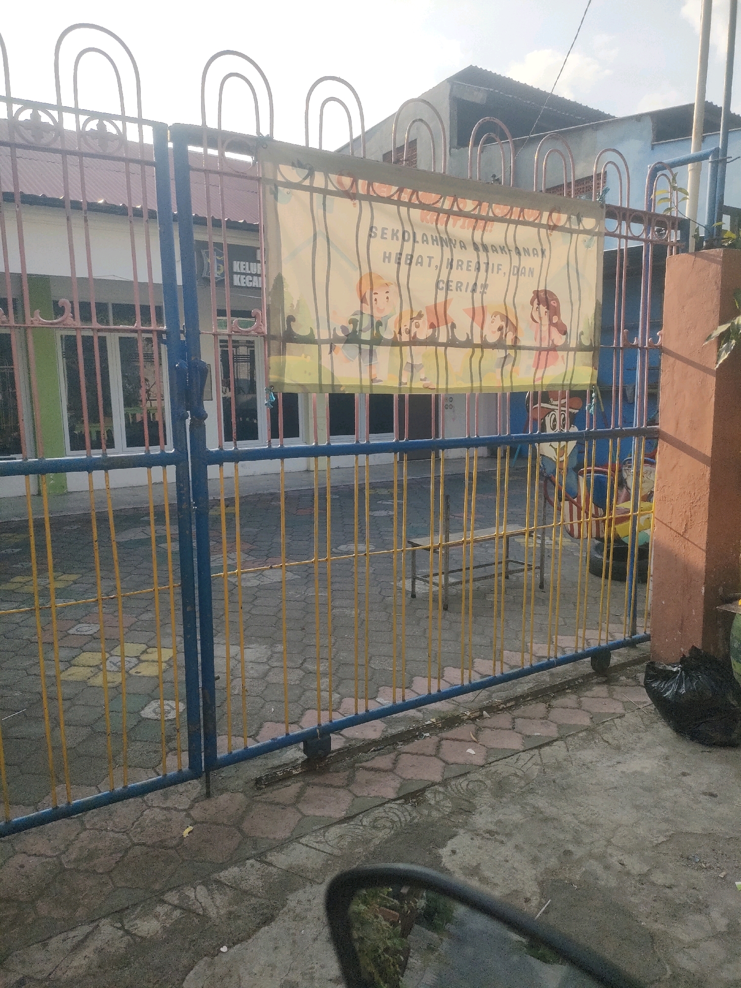 The image shows a chain-link fence in front of a building that appears to be a school. A banner is hanging on the fence with cartoon characters and text in Indonesian that reads "SEKOLAHNYA ANAK-ANAK HEBAT, KREATIF, DAN CERIA" which translates to "THE SCHOOL OF GREAT, CREATIVE, AND CHEERFUL CHILDREN". The building behind the fence has windows and a green wall. The ground in front of the fence is paved with hexagonal tiles. A black trash bag is visible on the right side of the image. The lighting suggests it is daytime, with the sun casting shadows. The environment appears to be outdoors, possibly a schoolyard or entrance.