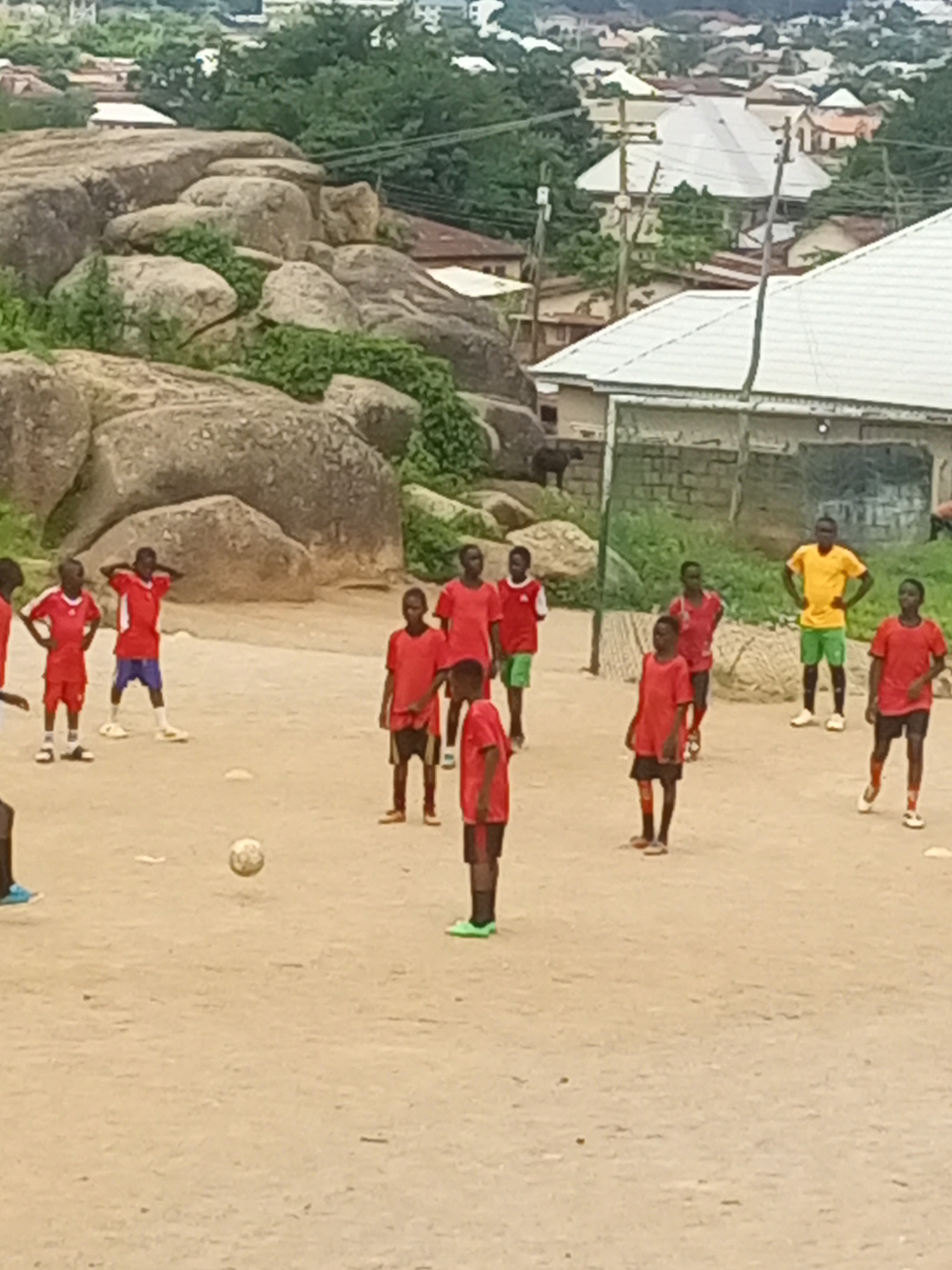 The image depicts a group of boys playing soccer in a dirt field, likely in Jos, Nigeria, given the distinctive rocky landscape that is characteristic of the region. In the background, the cityscape of Jos is visible with its characteristic red-tiled roofs, interspersed with lush greenery and large boulders. Several young boys, dressed in brightly colored soccer attire, are engaged in the game. One boy in the foreground is kicking a soccer ball. The scene suggests a casual game played outdoors in a community setting.