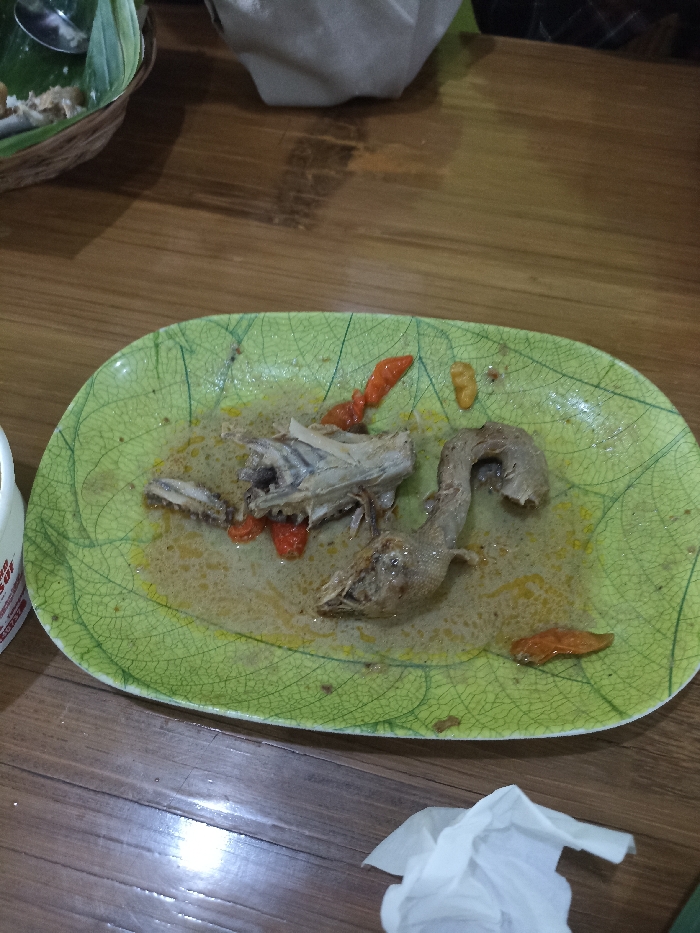 The image captures a close-up view of a meal's aftermath on a wooden table, likely in an indoor setting. The central focus is a green, rectangular plate with a distinctive leaf pattern and a cracked glaze texture. On the plate are remnants of a chicken dish, including several bones—a section resembling a chicken head or neck and part of a rib cage—submerged in a yellowish, creamy sauce or broth. Scattered among the bones and sauce are several bright red chili peppers, indicating a spicy dish.
To the upper left, partially visible, is a woven basket containing what appears to be discarded food scraps, possibly rice, along with a silver spoon. A crumpled white tissue or napkin rests on the wooden table surface in the bottom right corner. On the far left, the edge of a white container or cup is visible, displaying some unreadable red text.
The wooden table surface shows its natural grain and reflections, suggesting ambient indoor lighting. No people are visible, but the scene implies a recently concluded meal. The overall impression is of a table being cleared after dining. Given the context of Tulungagung, Indonesia, the dish could be a local chicken preparation common in Indonesian cuisine.