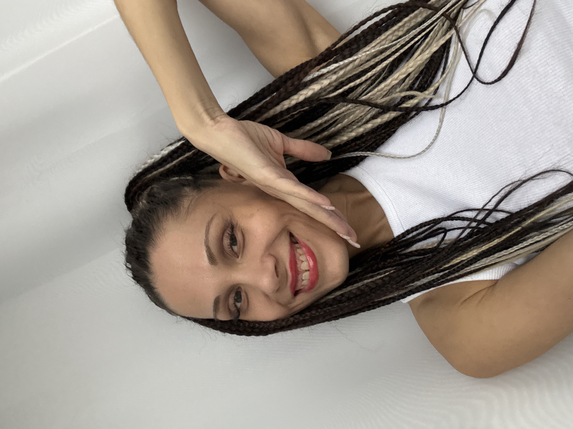 A woman with brown and blonde braids is smiling and partially covering her face with her right hand. She is wearing a white tank top. The background is a white, possibly indoor, setting. There is no indication of location in the city of Rusanovo, Russia.