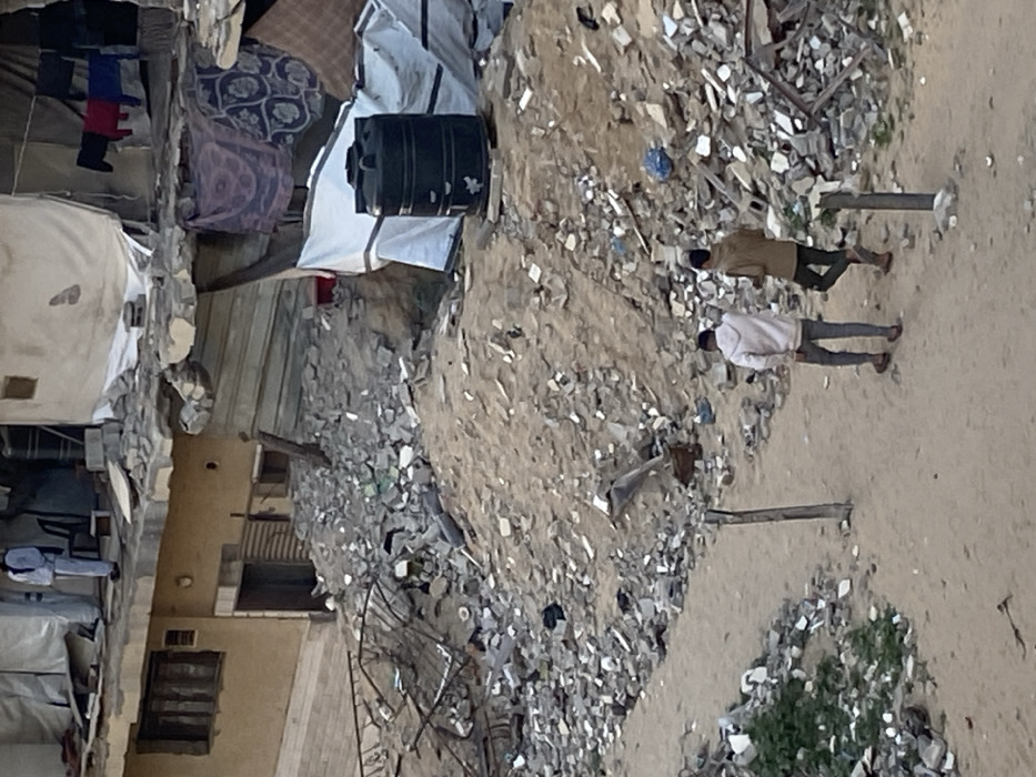 Two figures walk on a sandy path alongside debris. The buildings are heavily damaged, possibly due to conflict or natural disaster. Rubble, broken materials, and dust cover the scene. Clothes are hung outside near a water tank. Overall, the setting is impoverished and demonstrates evidence of hardship.