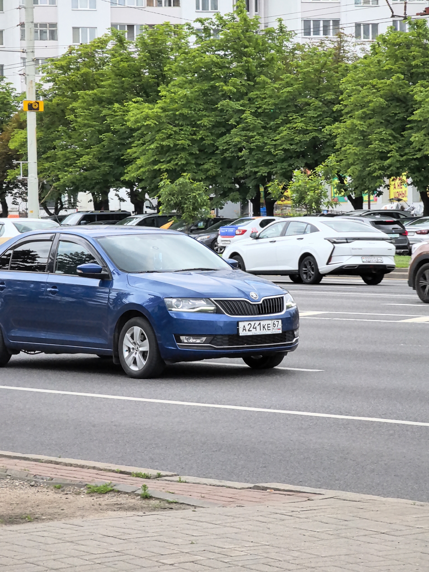 A blue sedan is parked on the right side of a street in a city. The car is facing right. The car is on the street, and there is a sidewalk to the right of the car. The sidewalk is made of gray concrete. There are trees and buildings behind the car. The buildings are tall and white. The trees are green and lush. There are other cars parked on the street. The sky is cloudy.