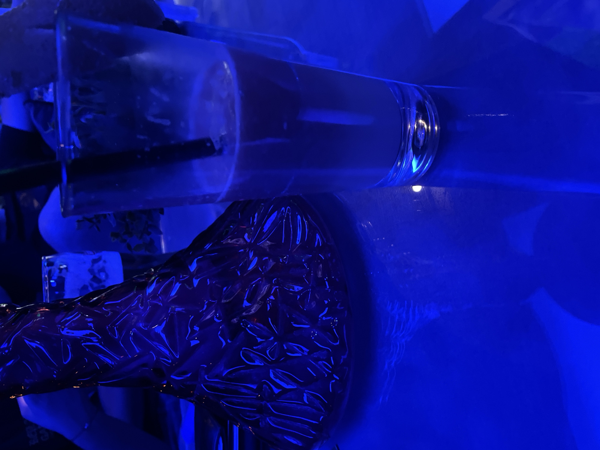 In an indoor setting with predominantly blue lighting, a glass containing a light brown beverage with a black straw stands on a reflective blue surface. To the left of the glass, a dark, textured object with a faceted surface is partially visible. The lighting creates a moody atmosphere.