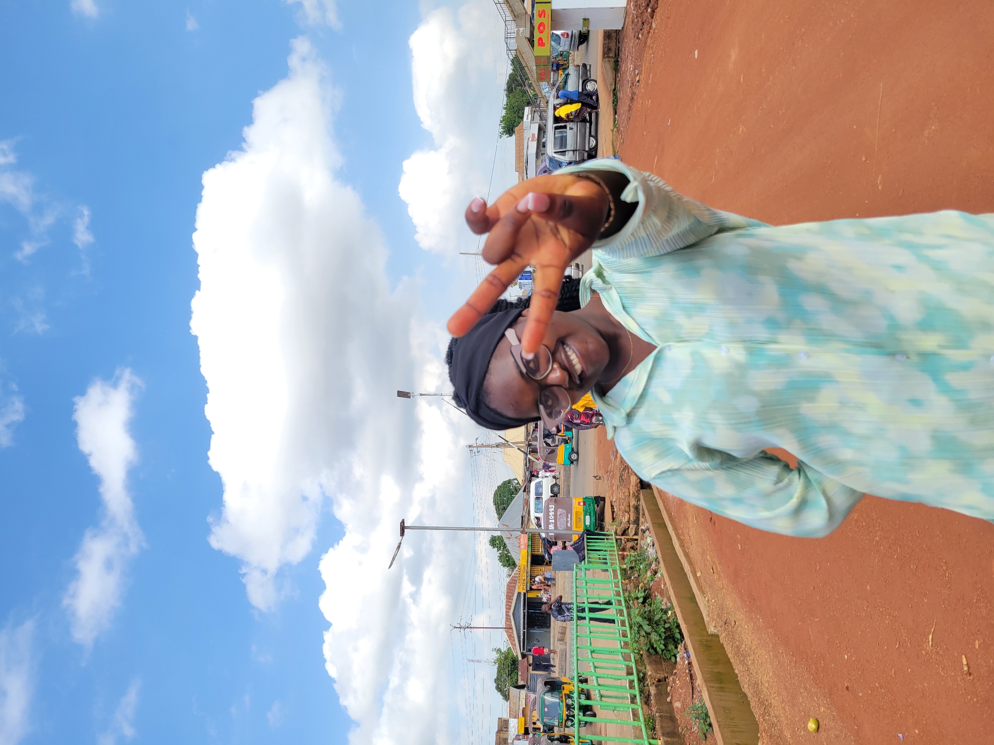 A young woman of color, wearing a light blue and green tie-dye patterned shirt and a black headscarf, smiles at the camera and holds up two fingers in a peace sign while standing on a dirt road in Gura Top, Nigeria. The background shows a bustling street scene with various vehicles, including three-wheeled auto-rickshaws and cars, as well as buildings and shops. A green metal fence lines the left side of the road, with overgrown vegetation visible behind it. The sky is a bright blue, dotted with fluffy white clouds. The overall impression is of a vibrant and lively urban environment.
