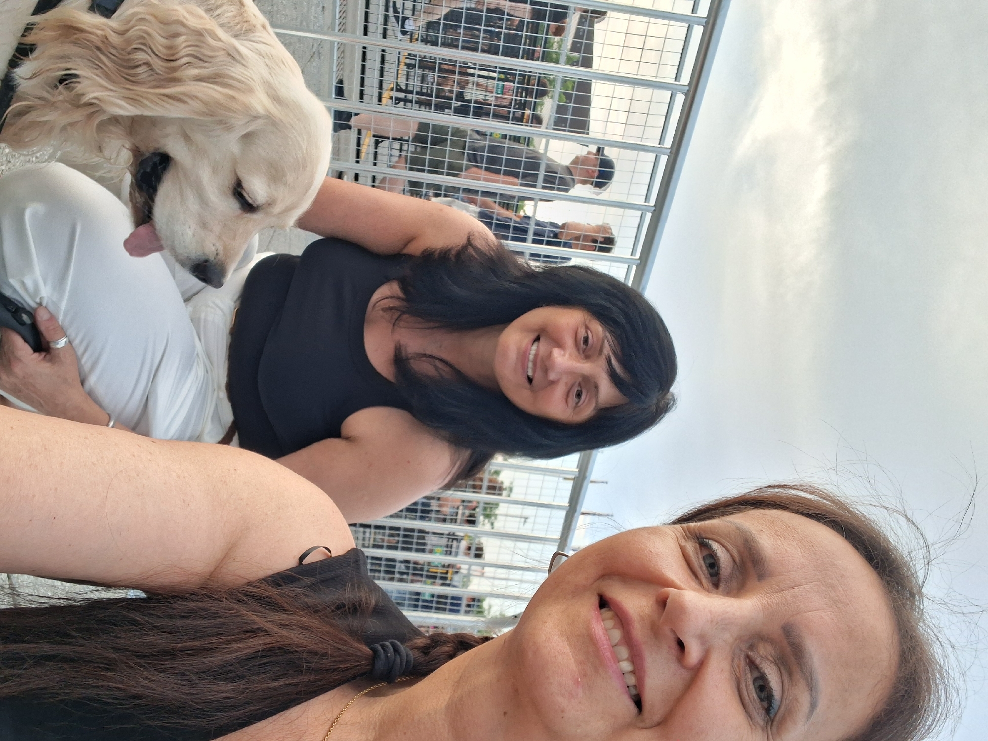 Two women and a golden retriever are posing for a selfie in an outdoor setting, possibly a public event or a concert, based on the crowd and metal barriers visible in the background. One woman is in the foreground, her face close to the camera, smiling. She has long, dark brown hair tied in a ponytail and is wearing a black tank top. The second woman is seated to her left, also smiling. She has dark hair and is wearing a black tank top and white pants. The golden retriever is in the lower left of the frame, looking to the side with its tongue slightly out. The background shows a metal fence with a crowd of people behind it, suggesting a ticketed event or a public gathering area. The sky is overcast with some light visible, indicating it could be late afternoon or early evening. The specific location in Ottawa, Canada, cannot be determined from the visual information alone.