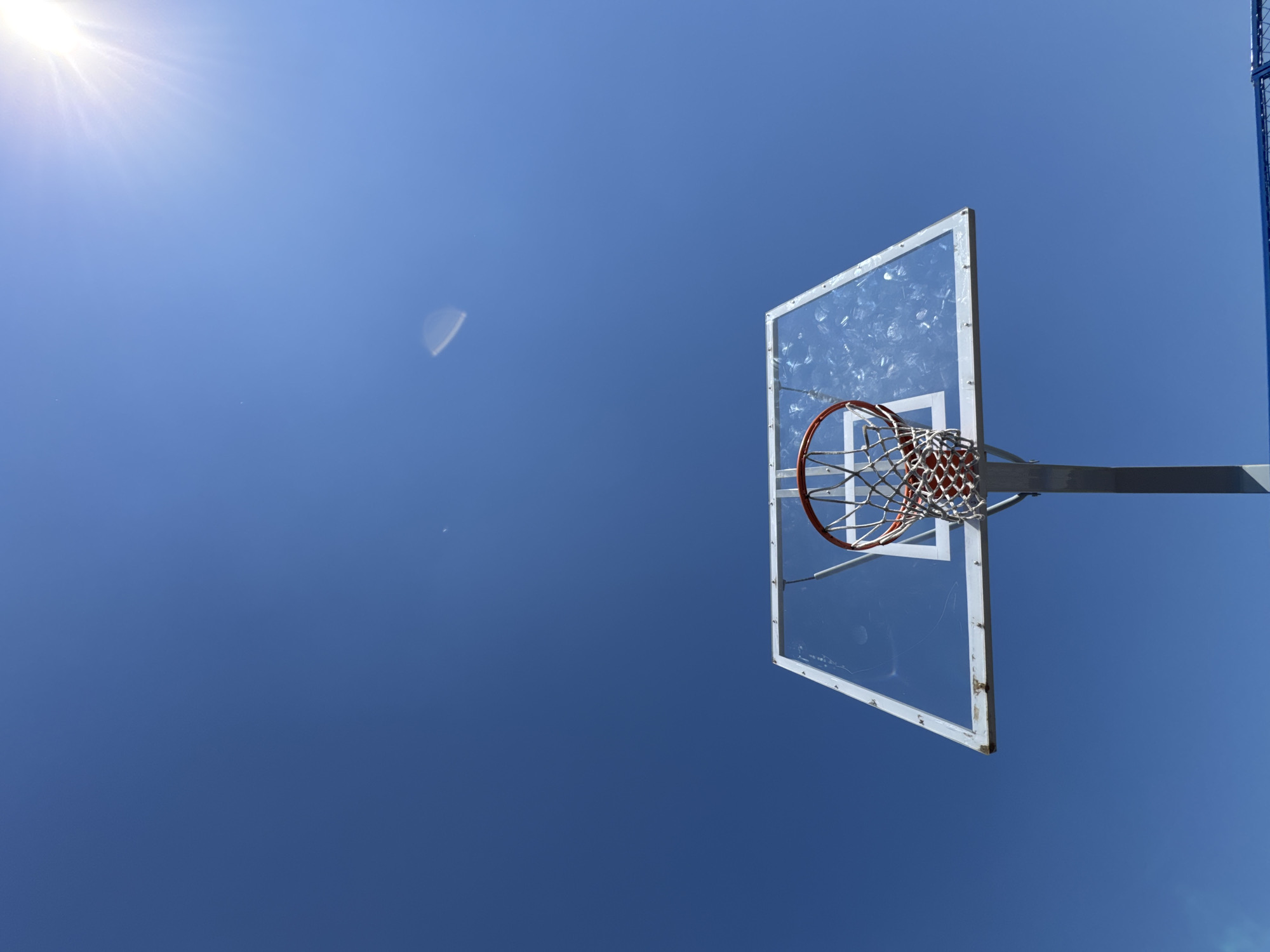 This upward-facing shot captures an outdoor basketball hoop set against a brilliant, clear blue sky. The sun, emitting bright rays, is visible in the upper left corner, creating some lens flare artifacts, including a distinct crescent shape further to the left in the sky.
The main subject is the basketball hoop, viewed from below. It features a transparent backboard, showing signs of wear such as scratches and marks. A white rectangular outline is painted on the backboard. The rim is a vibrant red-orange, and it holds a white net. The metallic grey support structure for the backboard is also visible. To the far right, a segment of a blue vertical pole, part of the basketball stand, can be seen, indicating the presence of a full court setup.
The scene depicts a sunny day, likely during morning or afternoon, perfect for outdoor activities. There are no people visible, nor is any specific activity taking place at the moment of capture, though the setting implies potential for basketball play. No discernible text is present in the image. The location context is Vysokiy, Russia.
