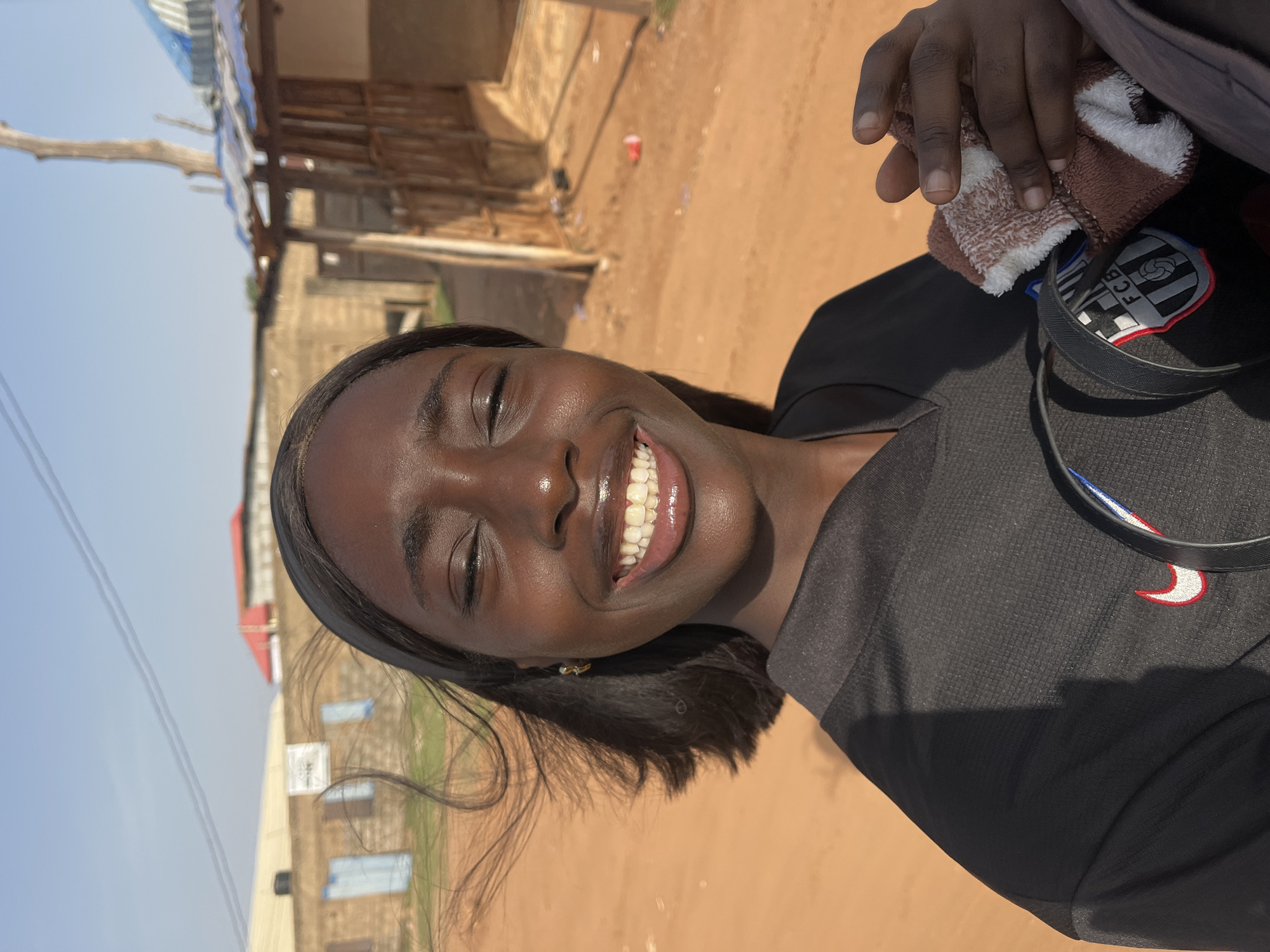 A young, dark-skinned woman smiles directly at the camera. She is wearing a black shirt with a white and red Nike swoosh on the shoulder. A headphone cable wraps around her neck with a patch visible with a possible logo. In her hand is a tan and white towel. Behind her is a dusty landscape with buildings in the background. Given the dirt terrain and building style, the location is presumed to be a less developed part of a Nigerian city. It's daytime, and the sunlight illuminates her face. Due to limited information within the image, it's impossible to confirm the exact location in Abuja, Nigeria.