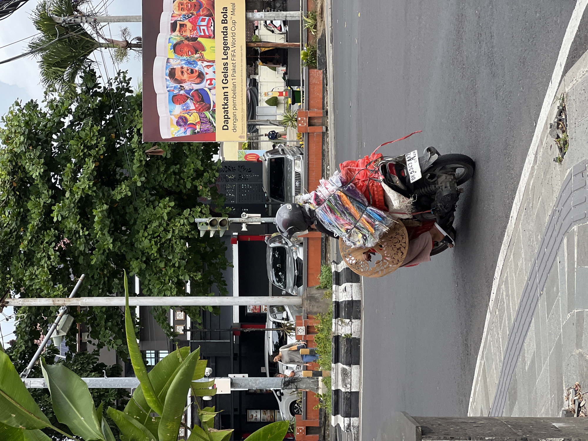 In Denpasar, Indonesia, a person on a scooter navigates a street. The scooter is laden with merchandise, including what appears to be umbrellas and other goods bundled together with red and clear plastic wrap, and a decorative wooden item secured to the rear. The driver is wearing a helmet and a pink garment. In the background, cars are visible on the road, along with a large billboard advertising a FIFA World Cup promotion featuring images of football players and cups. Trees and buildings line the street, indicating an urban environment. A pedestrian walkway with tactile paving runs along the side of the road, and a traffic light is visible. The scene appears to be a typical street view in a bustling city.