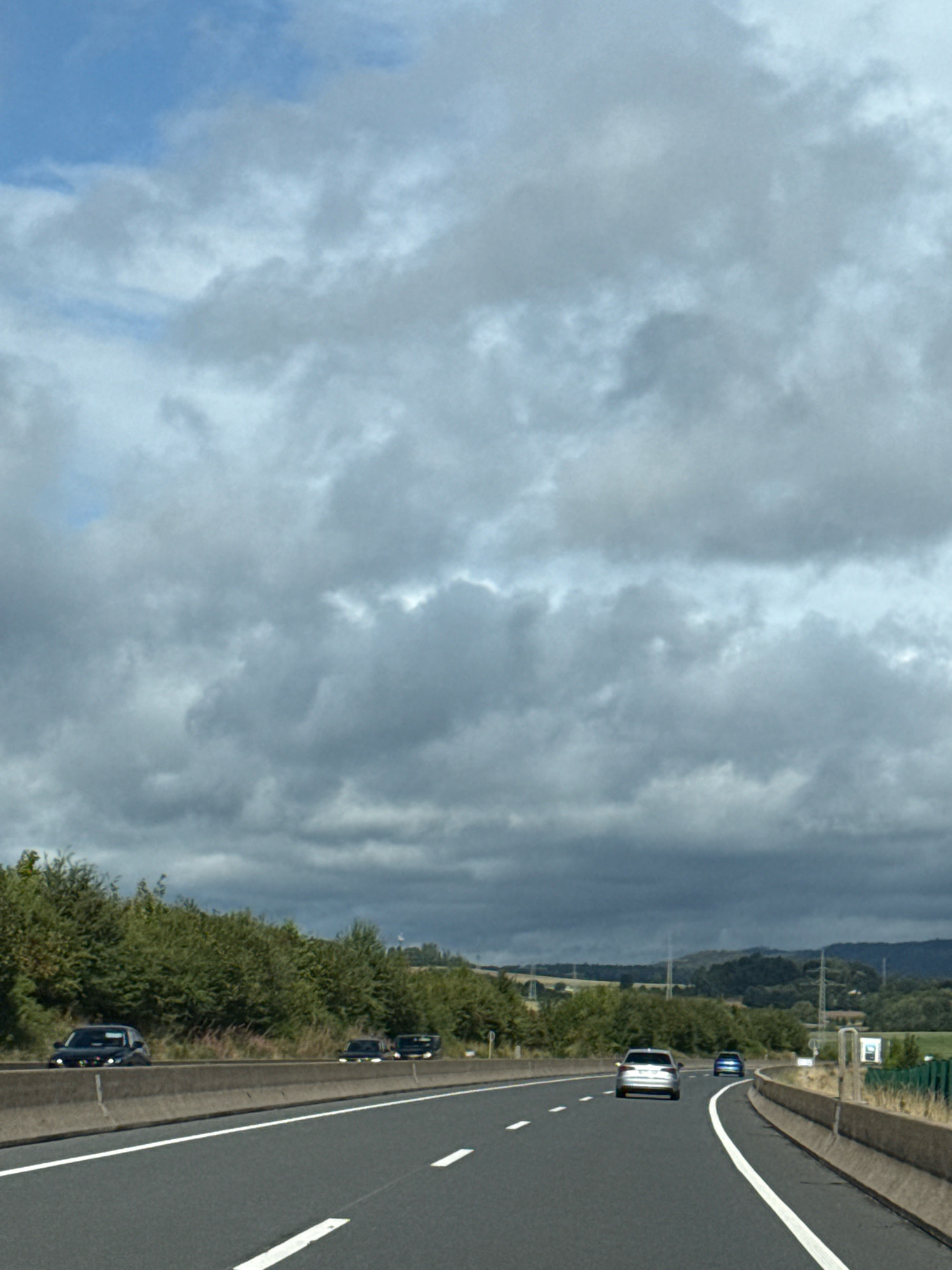 The image shows a view from a highway in Bad Sachsa, Germany. The highway has multiple lanes and is divided by a concrete barrier. There are several cars on the highway, including a black sedan in the left lane and a silver sedan and a blue sedan further ahead. The highway is surrounded by trees and vegetation. The sky is cloudy, with some patches of blue visible. The overall scene is one of travel and movement on a German highway.