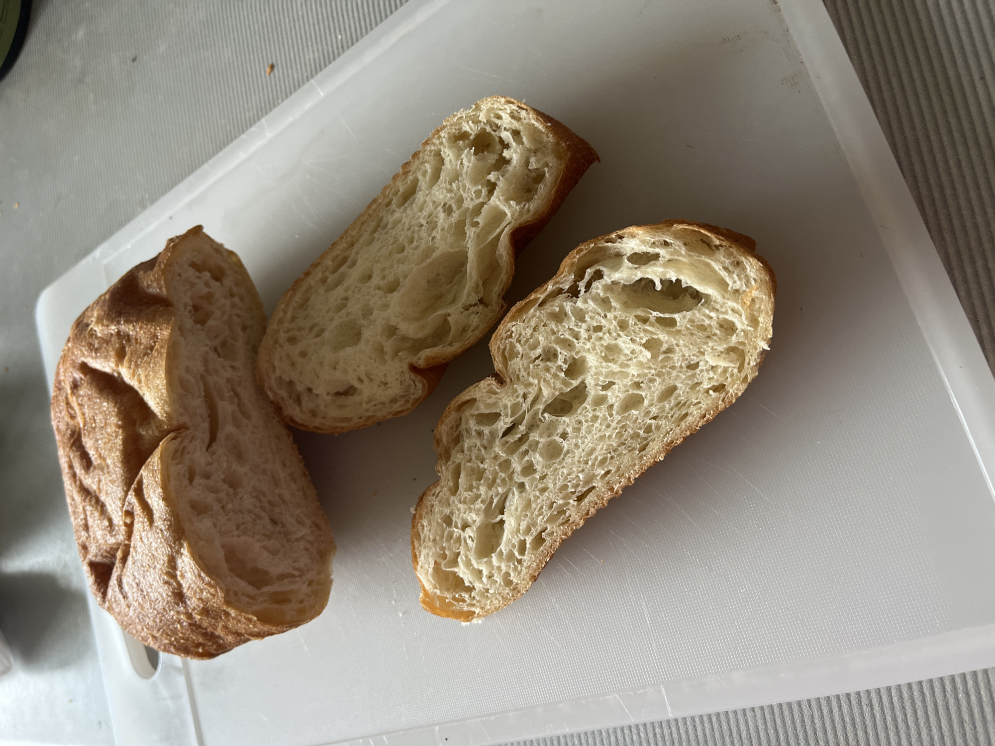 Two slices of bread, likely a baguette or similar artisan bread, are presented on a white cutting board. The bread slices have a golden-brown crust and an airy, irregular crumb structure with visible air pockets. One slice is whole, with its crust intact, while the other two appear to be cut pieces, revealing the soft interior. The cutting board has a subtle textured surface and is placed on a grey, ribbed surface, possibly a placemat or countertop. The lighting suggests it is daytime, and the overall scene is simple and focused on the food. There is no visible text in the image. The location context of Sochi, Russia, is provided but not directly inferable from the visual content of the image itself.