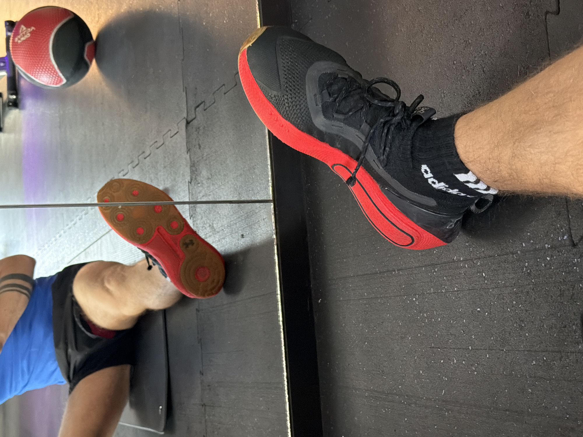 The image shows a person's legs and feet in a gym. One leg is extended forward with a red and black athletic shoe on the foot. This shoe has black laces and black socks with white stripes and logos. The other leg is bent, and the sole of a red and brown athletic shoe is visible. The person is sitting on a black exercise mat. In the background, reflected in a mirror, is a red and black medicine ball. The floor is made of dark gray interlocking mats. The scene appears to be indoors, possibly a fitness studio or gym. The image does not provide any information or context related to the city of Sofia, Bulgaria.