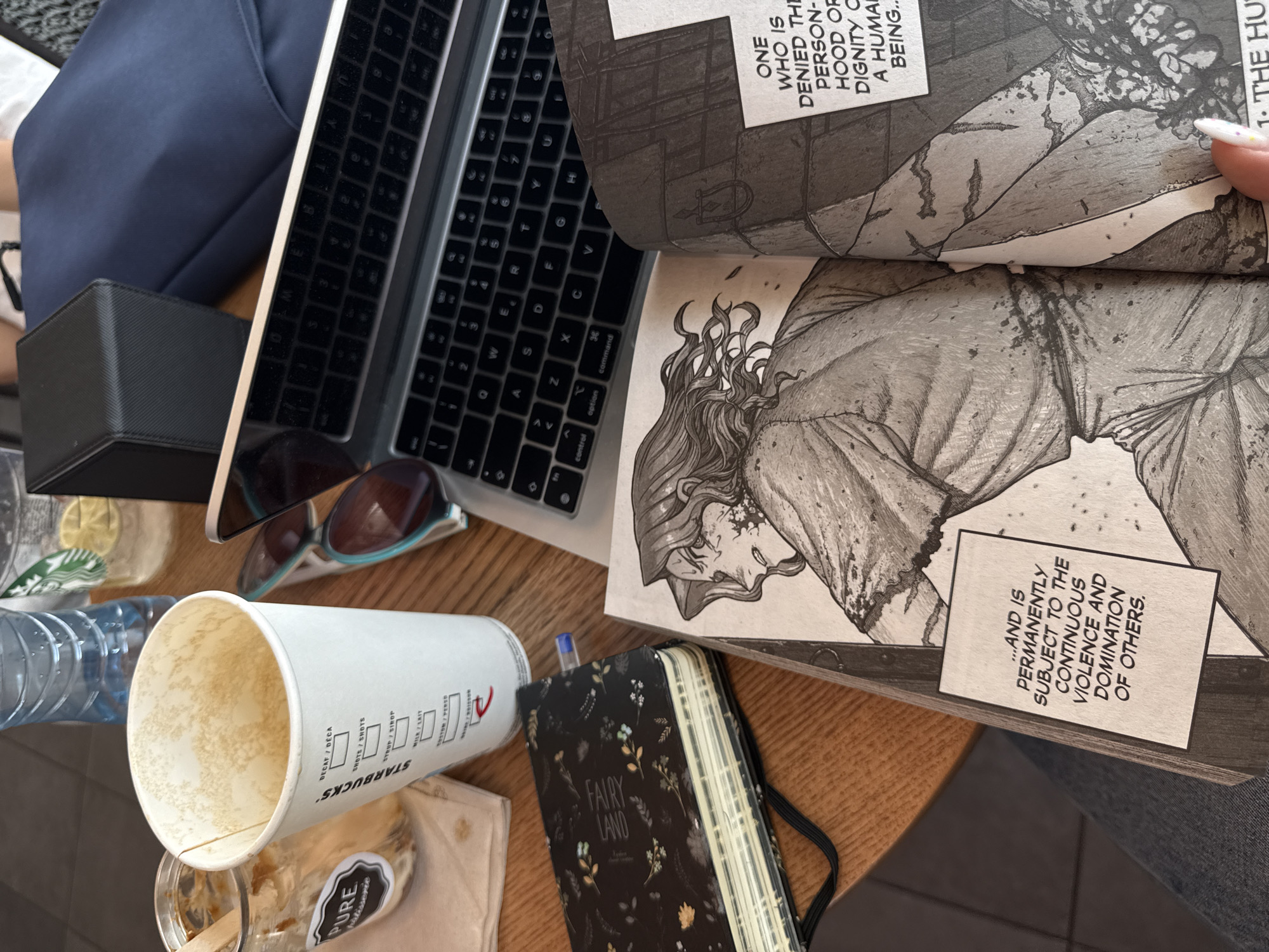 The image shows a close-up view of a wooden table with various objects, suggesting a casual work or study setting. A laptop is open, its keyboard visible, next to a dark blue fabric item, possibly a bag or cushion. A black case sits near the laptop.
Dominating the foreground is a white Starbucks paper cup, mostly empty, with some residue at the bottom. To the left, a clear plastic water bottle is visible, and behind it, a glass with a lemon slice and ice. A pair of sunglasses with light blue frames rests on the table, reflecting the surroundings.
Further into the image, a manga book is open, revealing black and white illustrations and text. One page shows a close-up of a person's head and shoulders with long, wavy hair, rendered in a detailed, somewhat somber style. Another section of the manga contains text in English, with phrases like "ONE WHO IS DENIED THE PERSON-HOOD OF DIGNITY" and "...AND IS PERMANENTLY SUBJECT TO THE CONTINUOUS VIOLENCE AND DOMINATION OF OTHERS."
A small black notebook with a floral pattern and the words "FAIRY LAND" on its cover is also present, with a blue pen inserted into its spine. The overall impression is one of a personal workspace, possibly during the day given the lighting, with items suggesting a break from work or study. The location context of Sofia, Bulgaria, does not offer specific visual cues within the image itself.