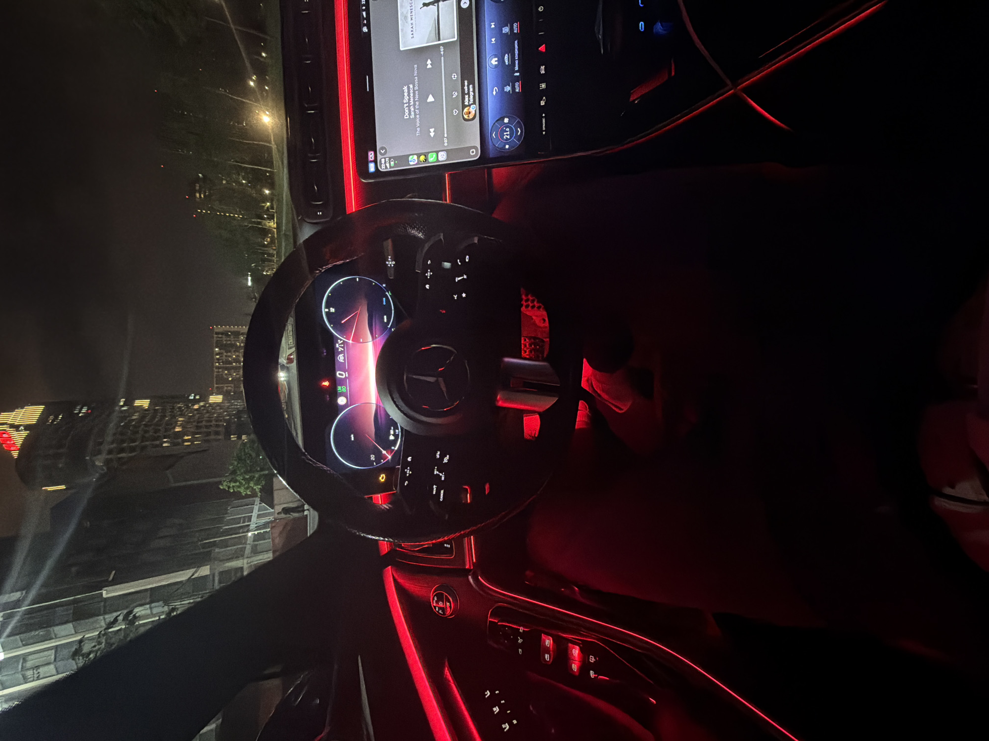 The image, taken inside a car at night in Moscow, Russia, shows a view of a city skyline through the windshield. The car’s interior is illuminated by red ambient lighting, highlighting the dashboard, steering wheel, and center console. The car's infotainment system displays a music track titled "Don't Speak" by Sarah Menescal. The cityscape visible through the windshield features various lit buildings, indicating an urban environment.