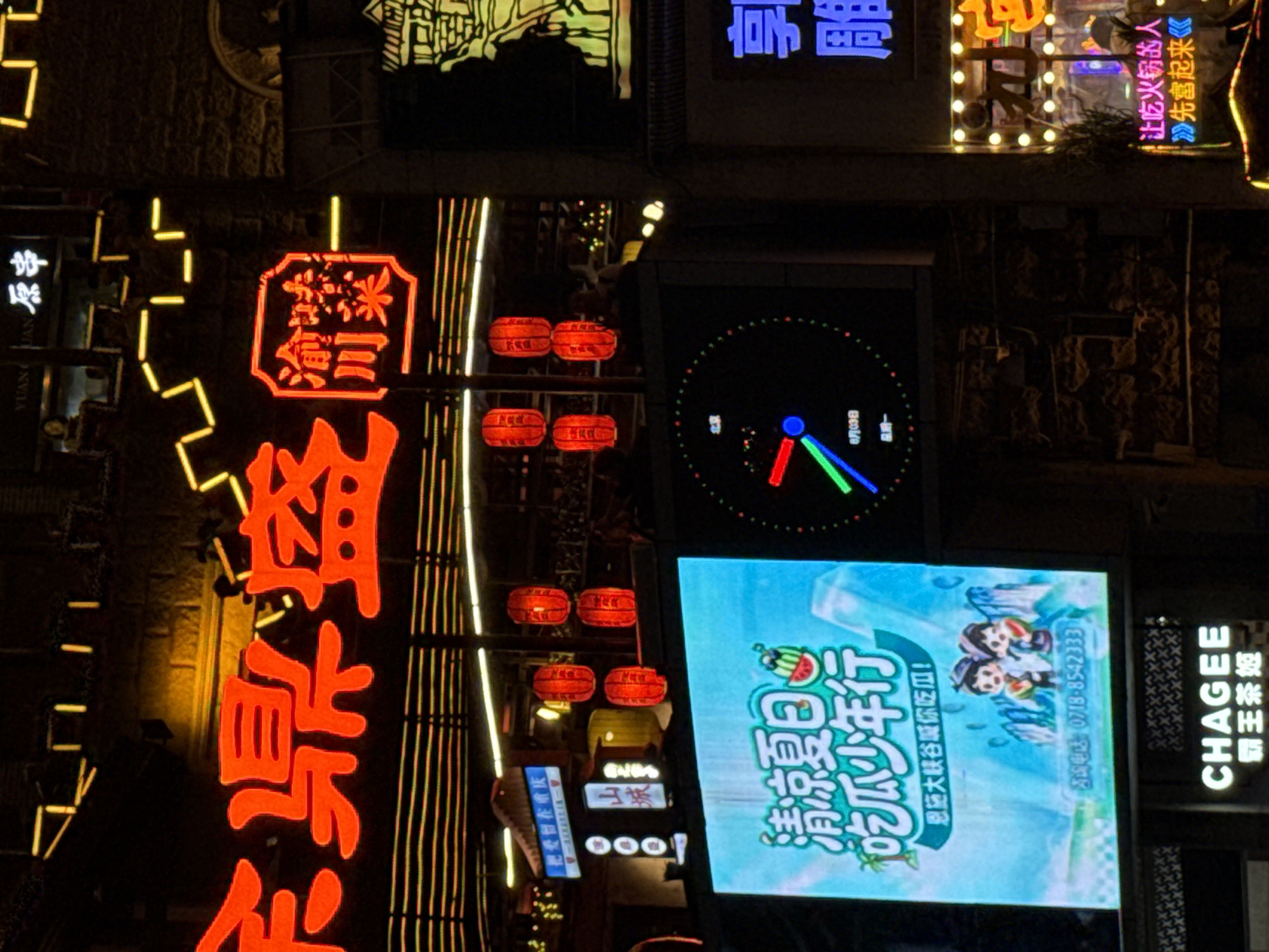 The image shows a nighttime street scene in Yuzhong District, China, likely a commercial area with illuminated signage. Dominating the upper portion of the image is a large red neon sign with Chinese characters, likely advertising a business. To the right of this, a smaller square sign with red characters within a red border is visible, also in neon. Below the large red sign, a display screen shows vibrant graphics, including cartoon figures and watermelon imagery, with Chinese text, and a phone number. To the right of this screen, a digital clock with colorful hands and markings is displayed. Further to the right, a bright blue neon sign with Chinese characters is present. The background includes brick walls and architectural details, some of which are outlined with yellow neon lights. Several red lanterns are suspended in the scene, adding to the festive nighttime atmosphere. The overall impression is one of a bustling urban environment after dark, with businesses using prominent neon signage to attract customers.