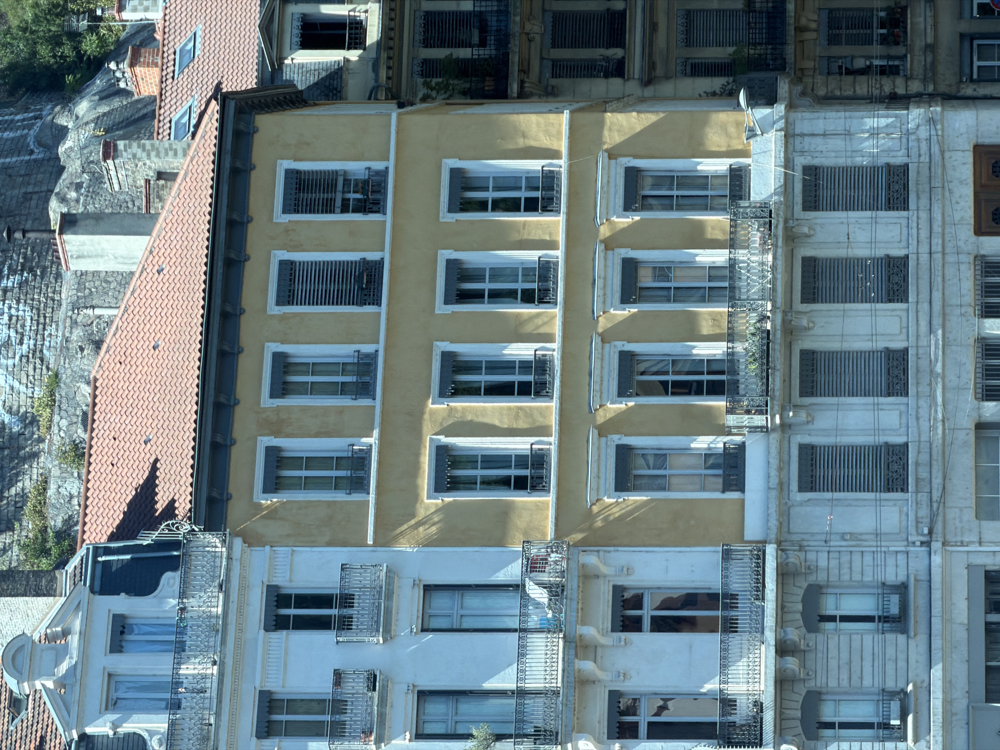 The image shows a close-up view of the facades of several adjoining apartment buildings in Lyon, France, viewed from below and at a slight angle. The buildings have a classical architectural style, with multiple stories and many windows. The building on the left is painted white and has ornate balconies. The central building is painted yellow and has simpler window frames. The building on the right appears to be older, with darker stone or stucco. The rooftops are visible, covered with red tiles. Behind the buildings, a steep, rocky hillside is visible, partially covered with greenery and stone structures. The lighting suggests it is daytime, with sunlight casting shadows on the facades. No people are visible in the image. The overall impression is of a dense urban environment with historical architecture.