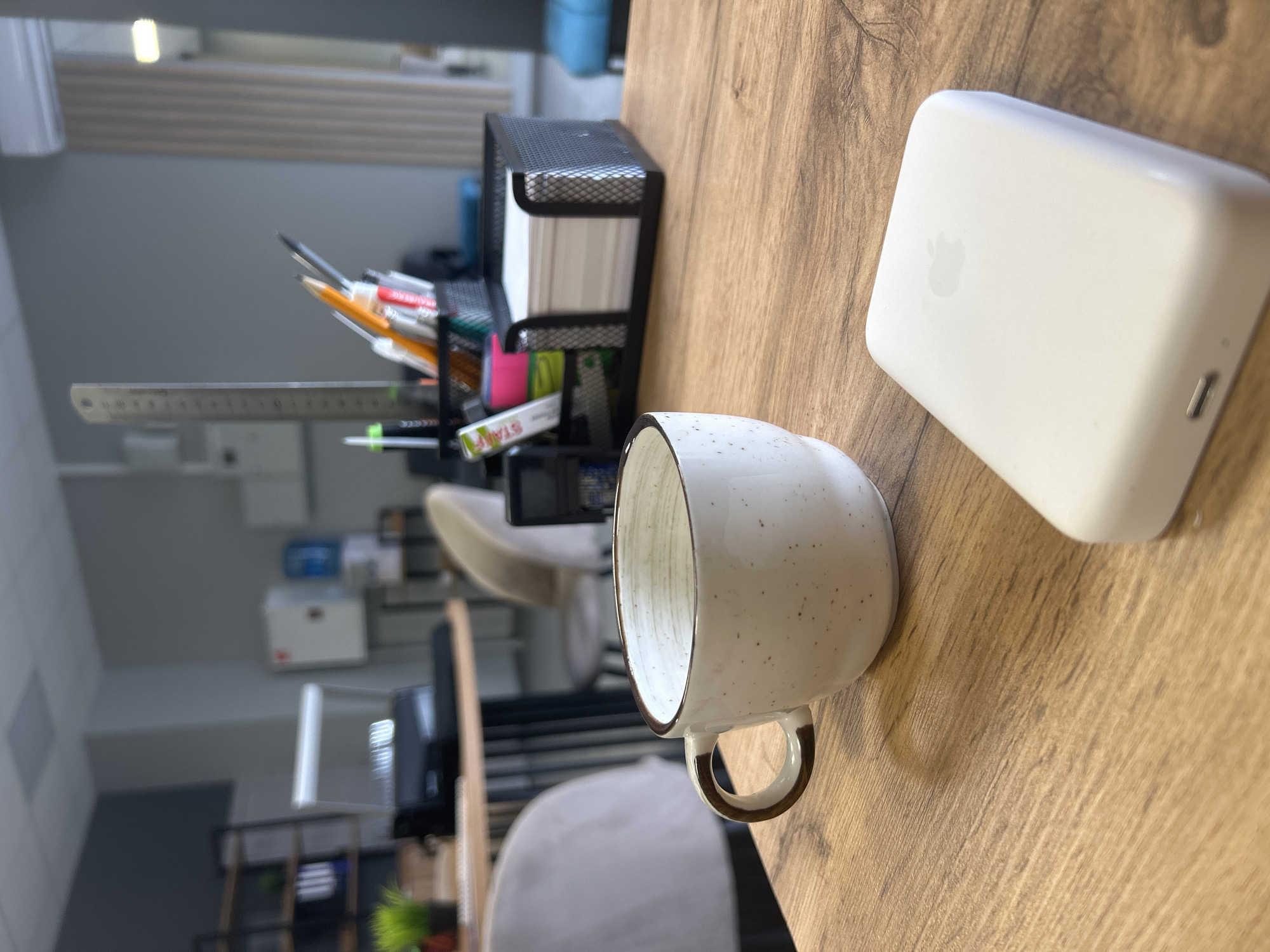 The scene depicts an interior office environment, presented as being located in the city of Izhevsk, Russia. The primary focus is on a light brown wooden desk surface.
In the foreground, an empty, speckled light-colored ceramic mug with a brown rim and handle rests on the desk. To its right, a white rectangular electronic device, identified by an Apple logo on its top surface and a visible port on one end, is also on the desk.
In the midground, a black metal mesh desk organizer holds various writing implements including pens, pencils, highlighters, and a tall silver ruler. Adjacent to this, another black metal mesh container holds a stack of white square sticky notes.
The background reveals a modern office setting. To the left, a round table with a dark base is partially visible, flanked by two light-colored upholstered chairs. Behind this table, a black metal shelving unit displays binders and other items, with a small green potted plant on an unseen surface. The walls are predominantly light grey, with a darker grey accent section to the left. Further back, a white wall-mounted cabinet and a blue water cooler dispenser are visible. To the right, a white wall-mounted split-system air conditioner is present near light-colored vertical blinds or wall paneling. The ceiling features square recessed light fixtures. No individuals are present in the frame.