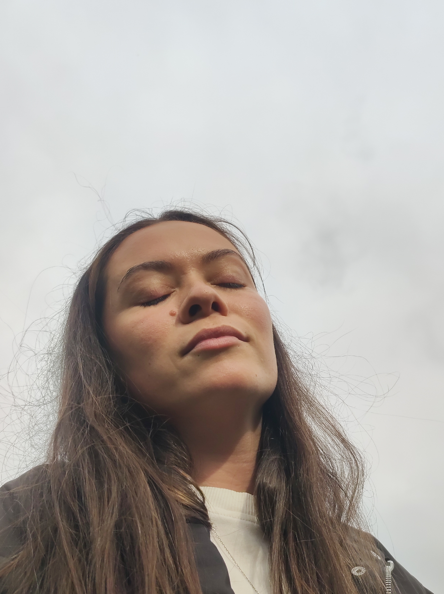 The image is a low-angle, close-up shot of a young woman's face against a cloudy sky. She has her eyes closed and her head tilted slightly upwards, appearing to be enjoying the outdoors. Her long, dark brown hair is windswept and frames her face. She is wearing a light-colored shirt and a dark jacket with some visible zippers and embellishments. A faint mole is visible on her right cheek. The background consists of an overcast sky. There is no discernible urban or natural environment present in the image to associate it with Chaikovsky, Russia.